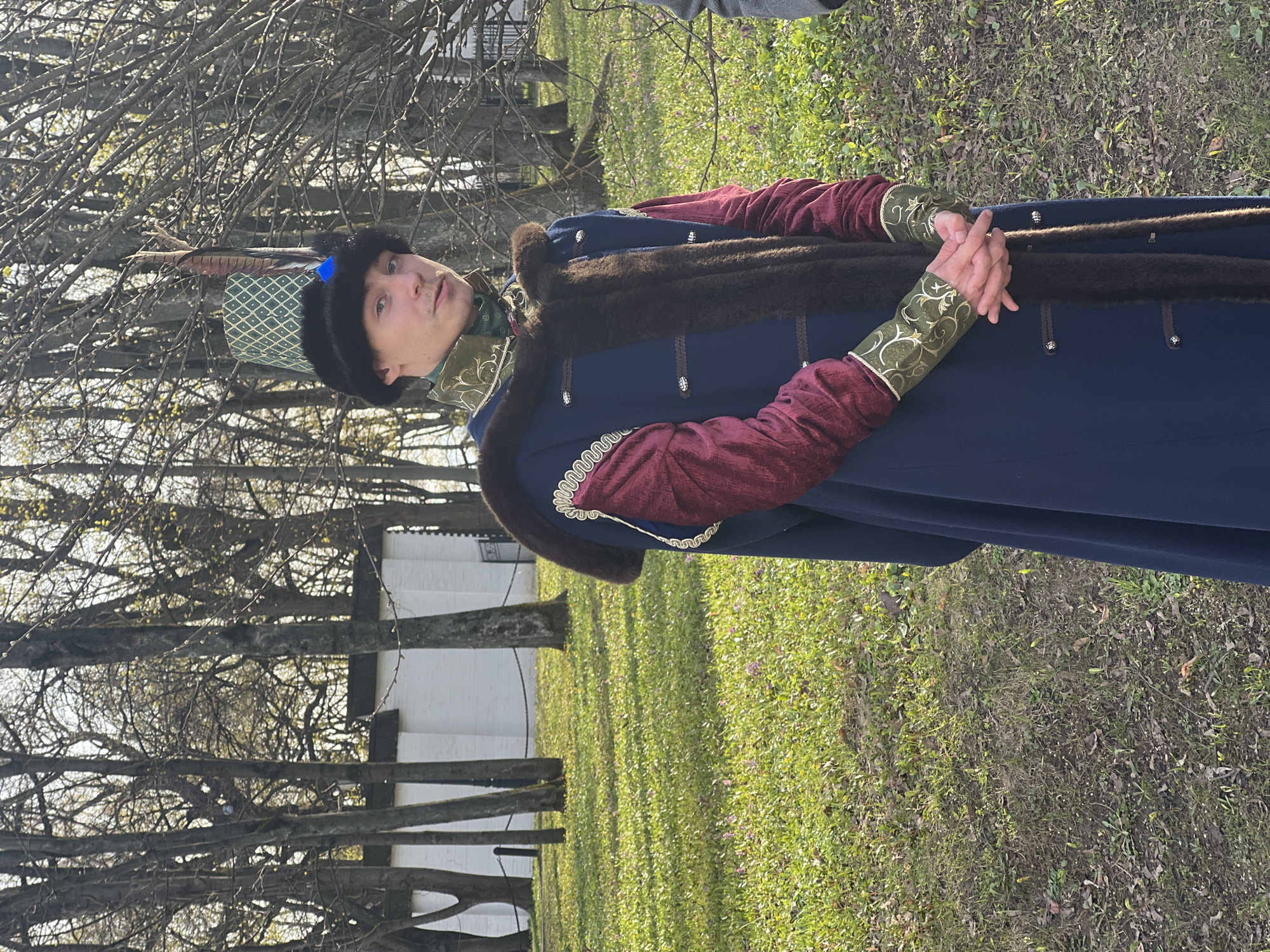 A medium-shot, eye-level photo shows a man standing on a grassy lawn. He is dressed in what appears to be a historical costume: a navy blue overcoat trimmed with dark brown fur, red sleeves with ornate gold cuffs, and a green and gold checkered hat with a feather. He has his hands clasped in front of him. The background includes trees with bare branches, a white building with a wooden fence, and other greenery. Given the attire and architectural style, it is plausible this photo was taken within the city of Moscow, Russia, potentially at a historical site or during a cultural event.