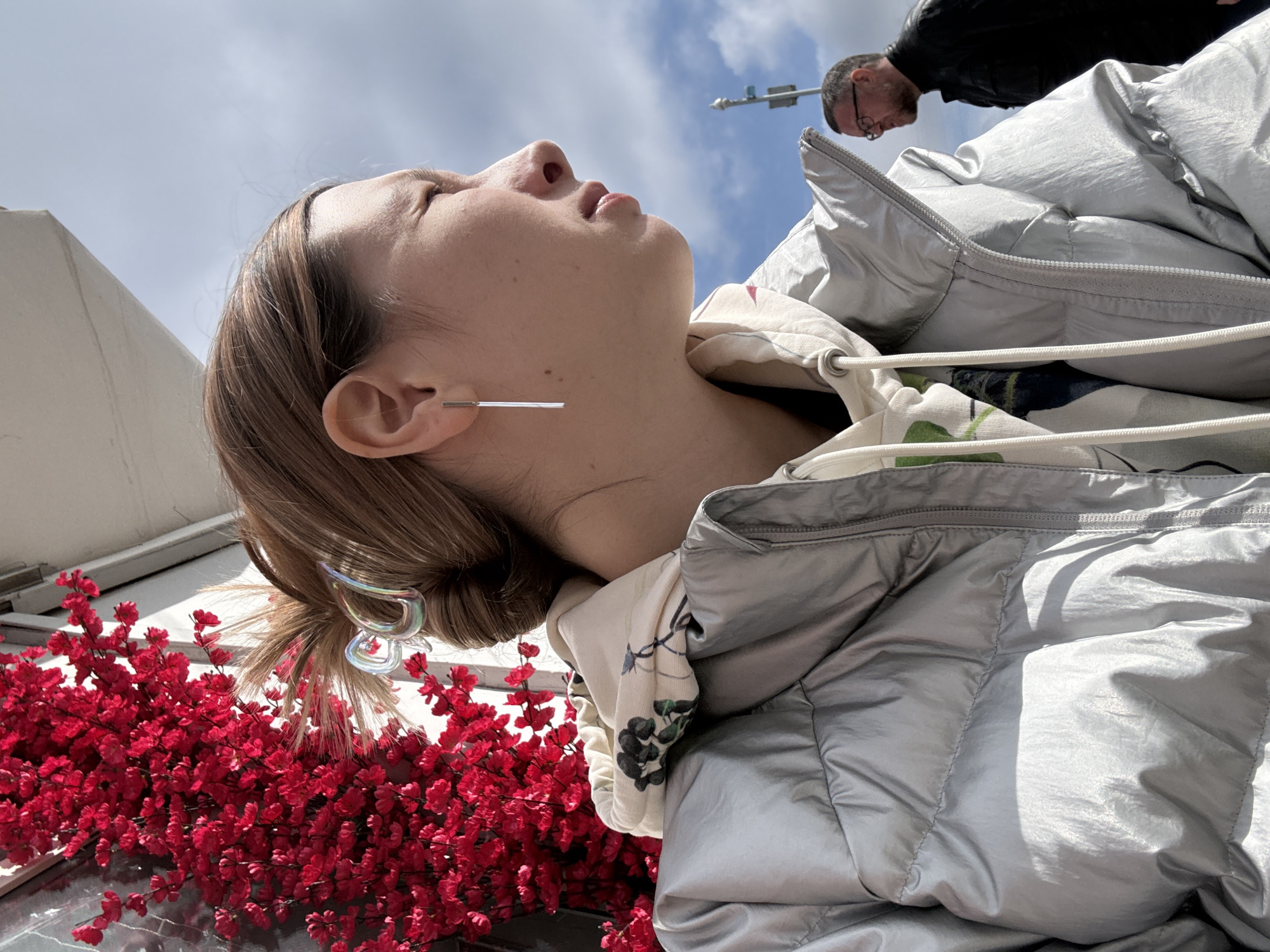 A woman wearing a silver puffer jacket and straight silver earrings is seen from a low angle. Her head is tilted back, and her eyes are closed. Her hair is brown and pulled back in a clip. In the background, a man wearing glasses and a black jacket is partially visible. A street sign is visible in the background, along with blue sky and white clouds. Red flowers are visible in the foreground. The location is Moscow, Russia.