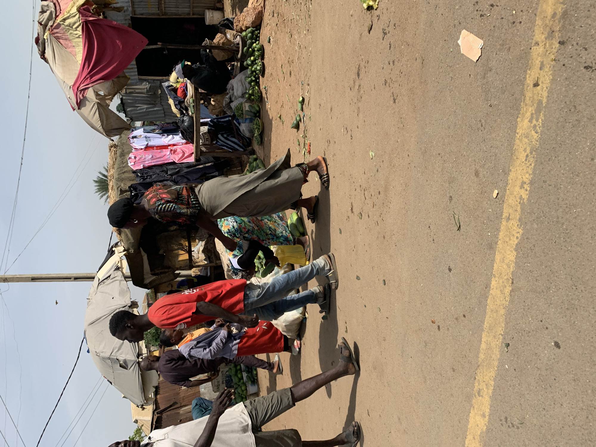 A daytime scene in what appears to be a roadside market in Rafin Par, Nigeria. Several individuals are present; some seem to be shopping while others are vendors. A woman wearing a head covering and a long skirt is handing a cucumber to a man in a red shirt and jeans. The market stall behind them is under makeshift awnings and displays merchandise, including clothes. There are piles of cucumbers, some on the ground near the yellow road line. The road is paved and slightly worn.