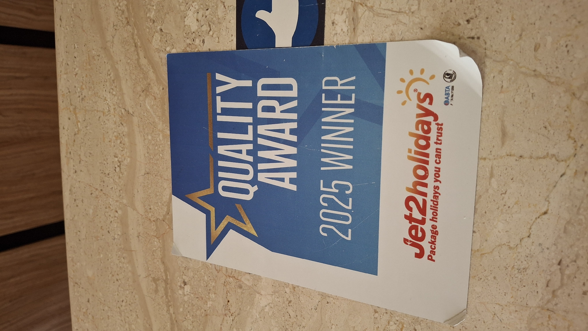 A Jet2holidays "Quality Award 2025 Winner" sticker is displayed on a light-colored marble countertop. The sticker is predominantly blue with a large golden star at the top left. "QUALITY AWARD" is written in large white capital letters, followed by "2025 WINNER" in white. Below this, the Jet2holidays logo appears in red with a sun graphic and the tagline "Package holidays you can trust". To the right of the Jet2holidays logo, there are two smaller circular logos, one labeled "ABTA" and another with an unknown symbol. The background of the sticker consists of a blue gradient. The environment appears to be indoors, with a wooden surface visible at the top of the image, and the countertop is speckled with brown and beige tones. There are no people visible in the image. The sticker is slightly creased on the right side, suggesting it might be attached to a surface or a brochure. No specific location in Nesebar, Bulgaria, can be identified from this image.