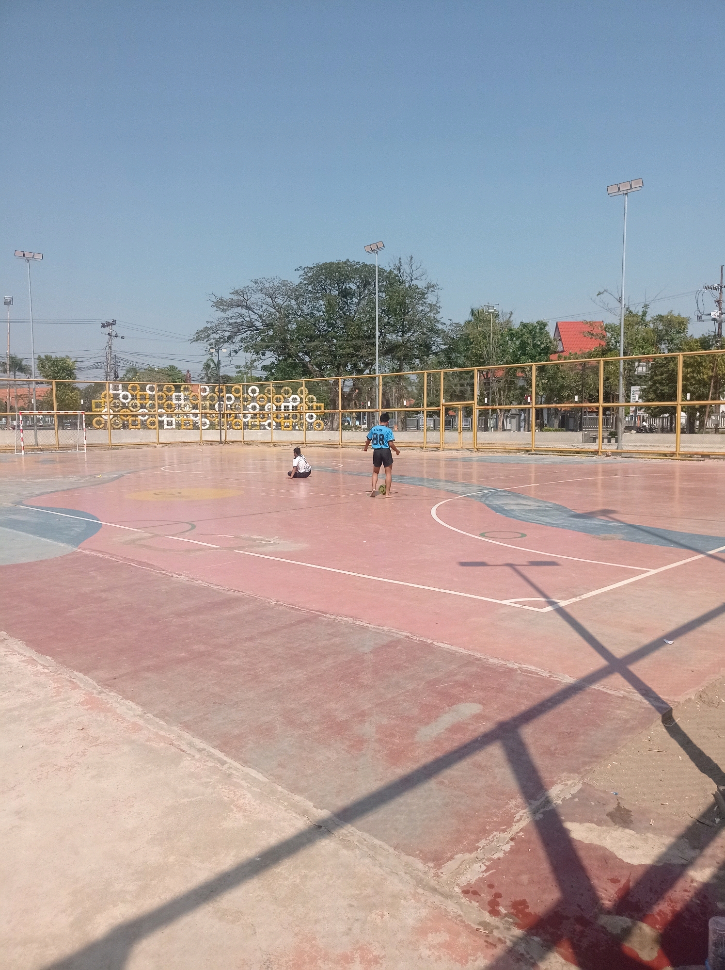 The image depicts an outdoor futsal court with a red and grey surface, marked with white lines indicating boundaries and areas. In the background, a yellow fence encloses the court, behind which are trees and buildings, including a house with a red roof. Tall lamp posts with multiple lights are visible, suggesting the court is equipped for evening play. Two young males are present on the court. One, wearing a blue jersey with the number "88" on the back and black shorts, stands near a green ball, seemingly about to play. The other, wearing a white and black outfit, is seated on the court. The sky is clear and blue, indicating a sunny day. The scene is likely located within a public recreational area in the city of Tuban, Indonesia, given the architectural style of the buildings and the tropical vegetation.