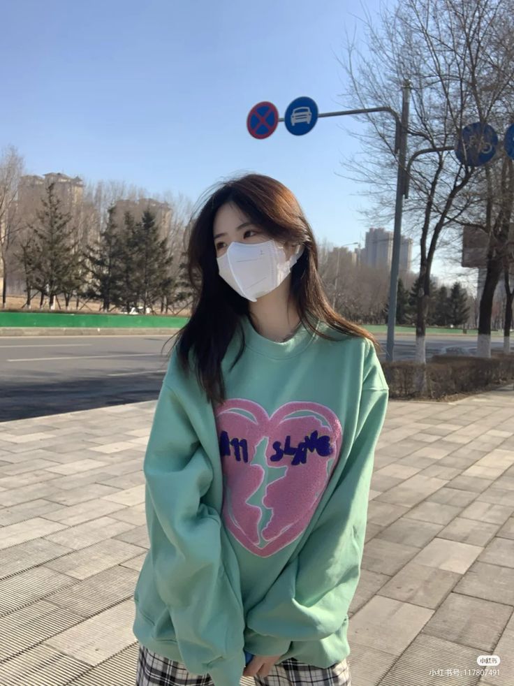 A young woman wearing a light green sweatshirt with a pink and purple heart design that says "in love" is standing on a sidewalk. The woman is wearing a white face mask. The sidewalk is paved with gray brick. There are trees lining the street and buildings in the distance. The street is empty and there are no cars present. There are two traffic signs on a metal pole behind the woman. The sign on the left is a red circle with a white cross inside. The sign on the right is a blue circle with a white silhouette of a car inside. The photo was taken in the city of Geneng, Indonesia, at the coordinates -7.45472 latitude and 111.427584 longitude. The scene is a typical urban environment. The photo was taken during the daytime. The weather is sunny and the sky is clear. The woman is looking to the right. The woman is standing on a sidewalk and is not interacting with anyone.