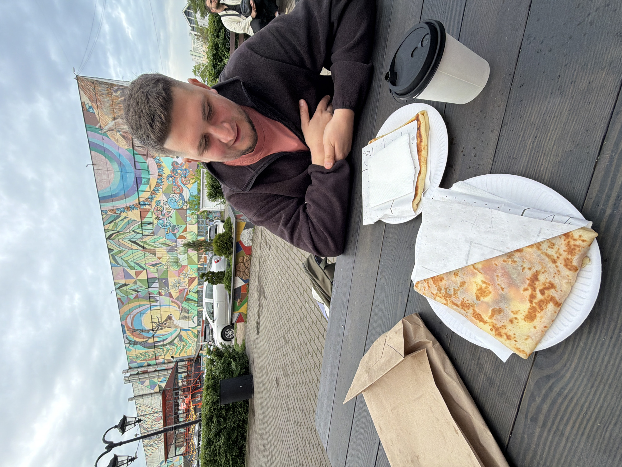 A man sits at a dark wooden table in an outdoor urban setting. Two crepes, wrapped in paper, sit on white plates, and a closed white coffee cup is to the right of them. To the left, a brown paper bag rests on the table. The man is wearing a dark jacket over a peach-colored shirt. Behind the table and the man is a view of a colorful mural on a building, with parked cars and greenery visible. The sky is overcast.