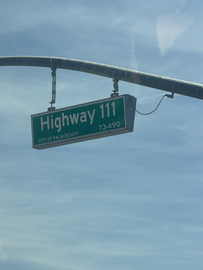 The image shows a green "Highway 111" sign with white lettering. Below that, it reads "CITY OF PALM DESERT" in smaller white lettering. The number range "73-490" is on the same line as "Highway 111", towards the right side of the sign. The sign is suspended from a curved, metallic pole by metal brackets. A thin black wire extends from the right side of the sign. The background is a bright blue sky with some light white clouds.
