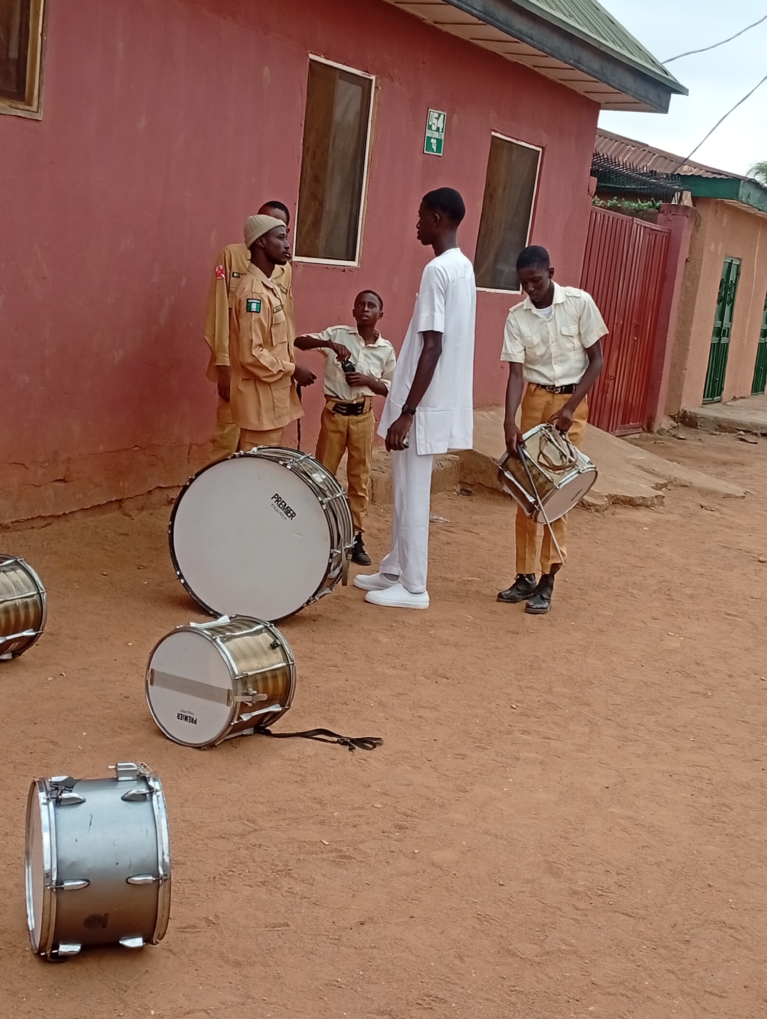 The scene is located outdoors in what appears to be a residential area of Narayi, Nigeria. The ground is a red-brown soil. There is a red building with several windows and a green house number sign displaying "54". A red metal gate is also visible.
Five young men are present. Two are wearing tan uniforms with hats and Nigerian flag patches on their shoulders. One is in white clothing from head to toe. The other two are wearing off-white shirts and tan pants.
Several drums are present. One large "PREMIER" branded bass drum is leaning against another drum. Two smaller drums are on the ground. One of the young men in tan pants is holding a small drum.
The young men appear to be interacting and preparing musical instruments, perhaps for a performance.