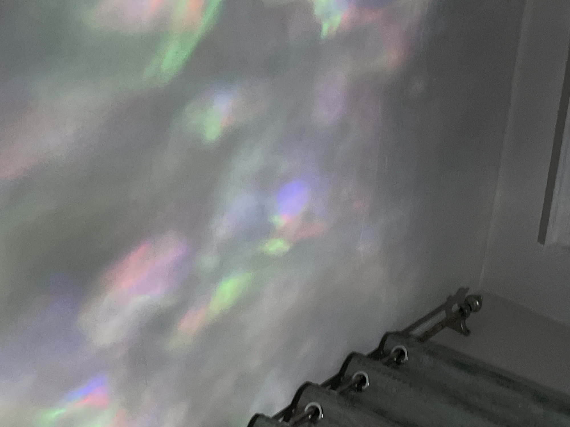 The image shows a ceiling with rainbow-colored light reflections. The reflections appear to be cast by a light source, possibly a projector or a prism, creating streaks and patches of color such as pink, purple, green, and blue. The ceiling is a light gray color and appears to have a slightly textured surface. In the lower left corner of the image, a portion of a gray curtain is visible, hanging from a curtain rod with metal grommets. The curtain is partially drawn, and a metallic hook or clip is attached to it. The overall scene suggests an indoor environment, likely a room in a residential or commercial building in Bishkek City, Kyrgyzstan. The lighting creates an abstract and colorful effect on the ceiling.