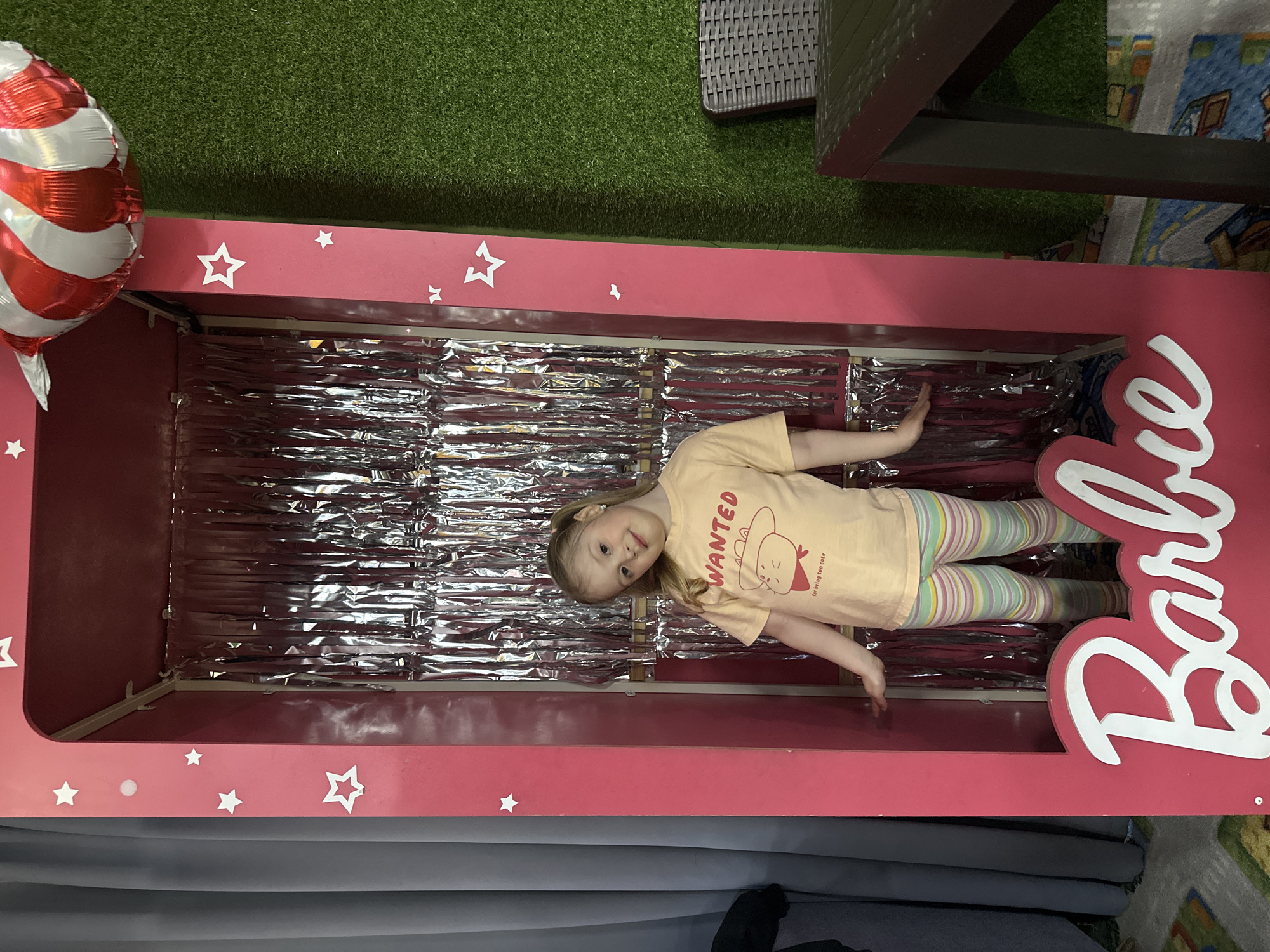 A young girl is lying inside a pink Barbie box. The box is decorated with white stars and the Barbie logo. The inside of the box is lined with reflective silver material. The girl is wearing a yellow shirt, and striped leggings. There is a red and white balloon to the side of the box. The setting appears to be indoors, with artificial grass and a table visible in the background. The city location is Glazov, Russia.