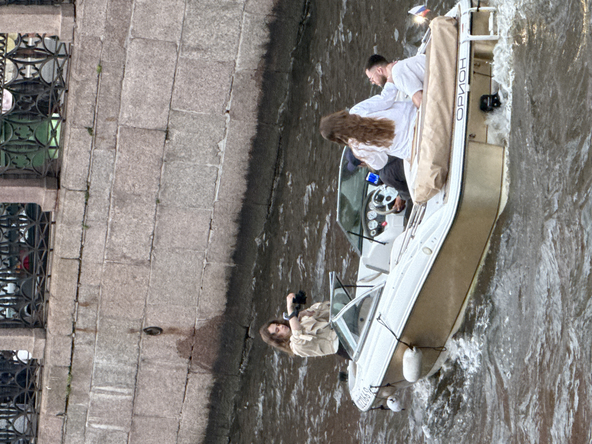 This media file shows a scene on a canal or river in Saint Petersburg, Russia. The main subjects are three people on a white motorboat. A man in a white shirt, wearing glasses and with short dark hair, is steering the boat. A woman with long, wavy brown hair, also in a white shirt, is seated next to him. In the foreground, another person, whose face is not clearly visible, is leaning over the side of the boat and holding a black camera, appearing to take a photograph.
The boat has "ОРИОН" written vertically on its side in black capital letters. The boat is moving, creating a wake in the water. The setting is a stone embankment, with what appears to be a decorative metal railing and windows visible on the left side of the frame. The overall lighting suggests it might be daytime, possibly overcast. The activity appears to be a leisurely boat ride, with the individuals enjoying the scenery and one person documenting the experience.
