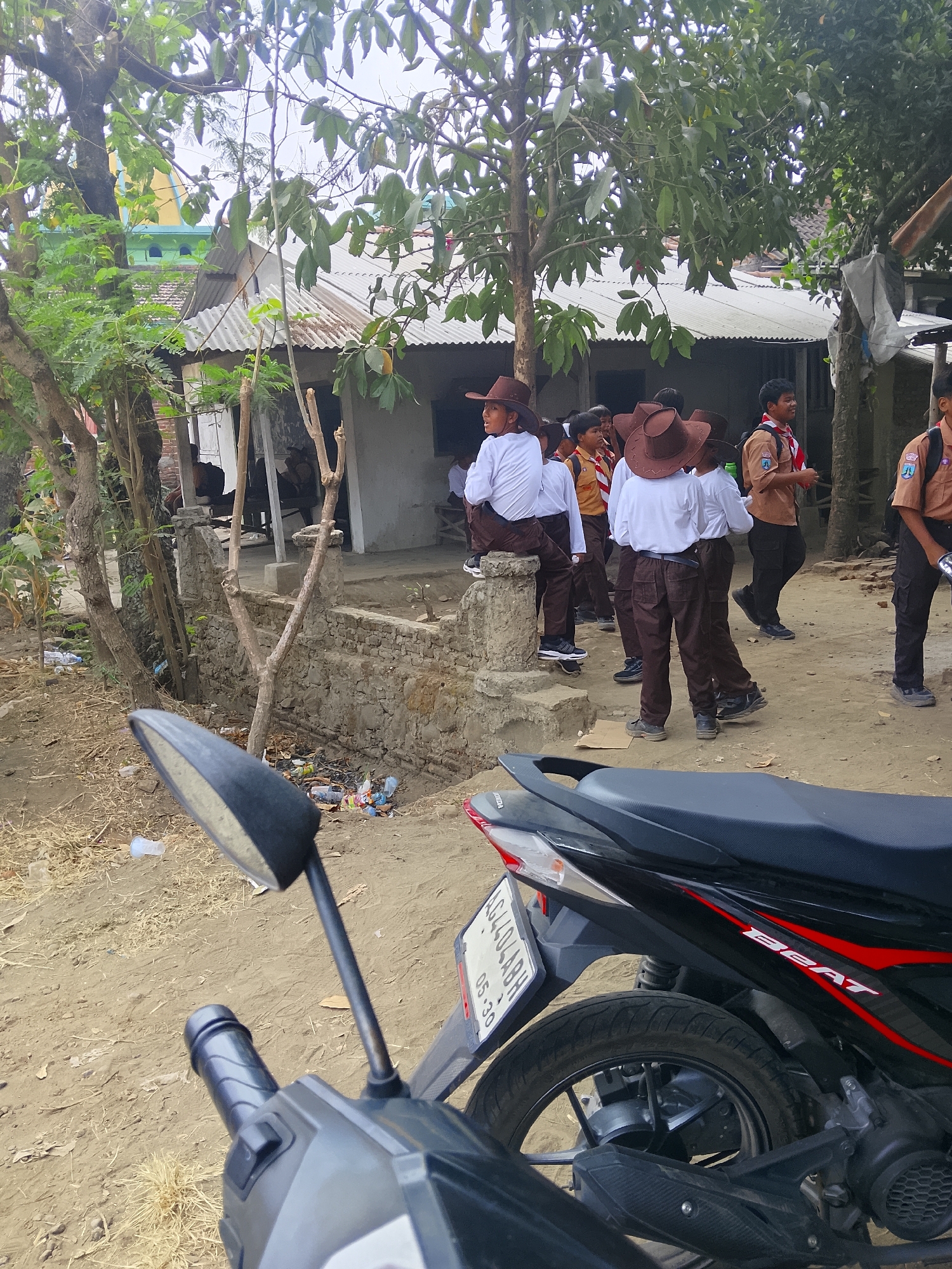 In Bangsri, Indonesia, a group of young Scouts in uniform, consisting of white shirts and brown pants with brown cowboy hats, are gathered in a dusty outdoor area. Some are seated on a stone wall and a concrete pillar, while others stand. A motorcycle is parked in the foreground, partially obscuring the lower portion of the image. Behind the Scouts, a building with a corrugated metal roof and a stone wall is visible, surrounded by trees and scattered debris, including plastic bottles and wrappers. The overall scene suggests a casual gathering or activity in a semi-rural or village setting.