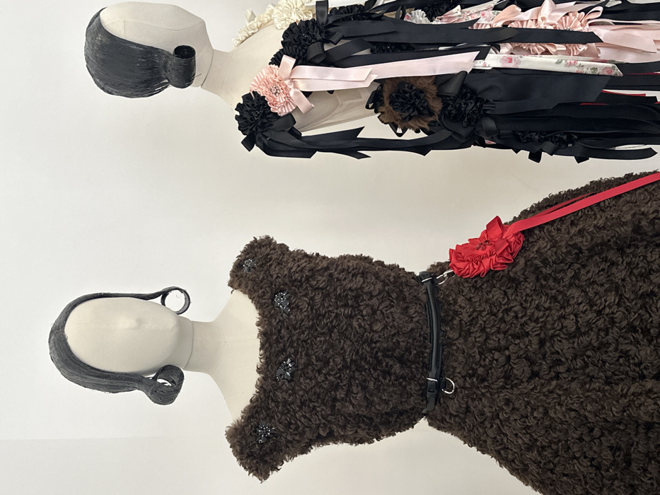 Two cream-colored, headless mannequins, each featuring a dark, stylized bob wig with side curls, are displayed against a plain white wall. The mannequin on the left wears a dark brown, curly faux fur sleeveless dress with a rounded neckline, embellished with three clusters of dark, reflective elements. A narrow black belt with two silver rings cinches its waist, from which a vibrant red ruffled fabric rosette with a long red ribbon hangs. The partially visible mannequin on the right exhibits a garment densely adorned with numerous black ribbons, interspersed with fabric rosettes and bows in black, light pink (some with subtle floral patterns), cream, and brown hues. The setting is an indoor exhibition space, likely a gallery or museum. The specific location within Paris, France, is not identifiable from the visual information provided.