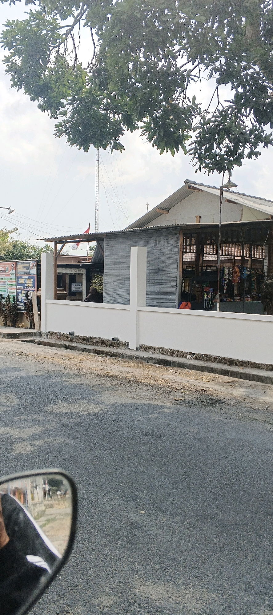 The image captures a street-level view in Bluri, Indonesia, dominated by a building with a corrugated metal roof, featuring an open-air section displaying various goods for sale. A white wall runs along the front of the property, with a paved road in the foreground. Overhead, tree branches with leaves and some unripe fruits are visible against a cloudy sky. A communication tower stands behind the building, and a small Indonesian flag is attached to the roof's overhang. A motorcycle mirror is visible in the lower left corner, reflecting a portion of the scene, including the building and trees. Two individuals are partially visible inside the building, one wearing an orange garment. A bulletin board with text and images is posted to the left of the building. The overall environment suggests a commercial or public space within a town or village.
