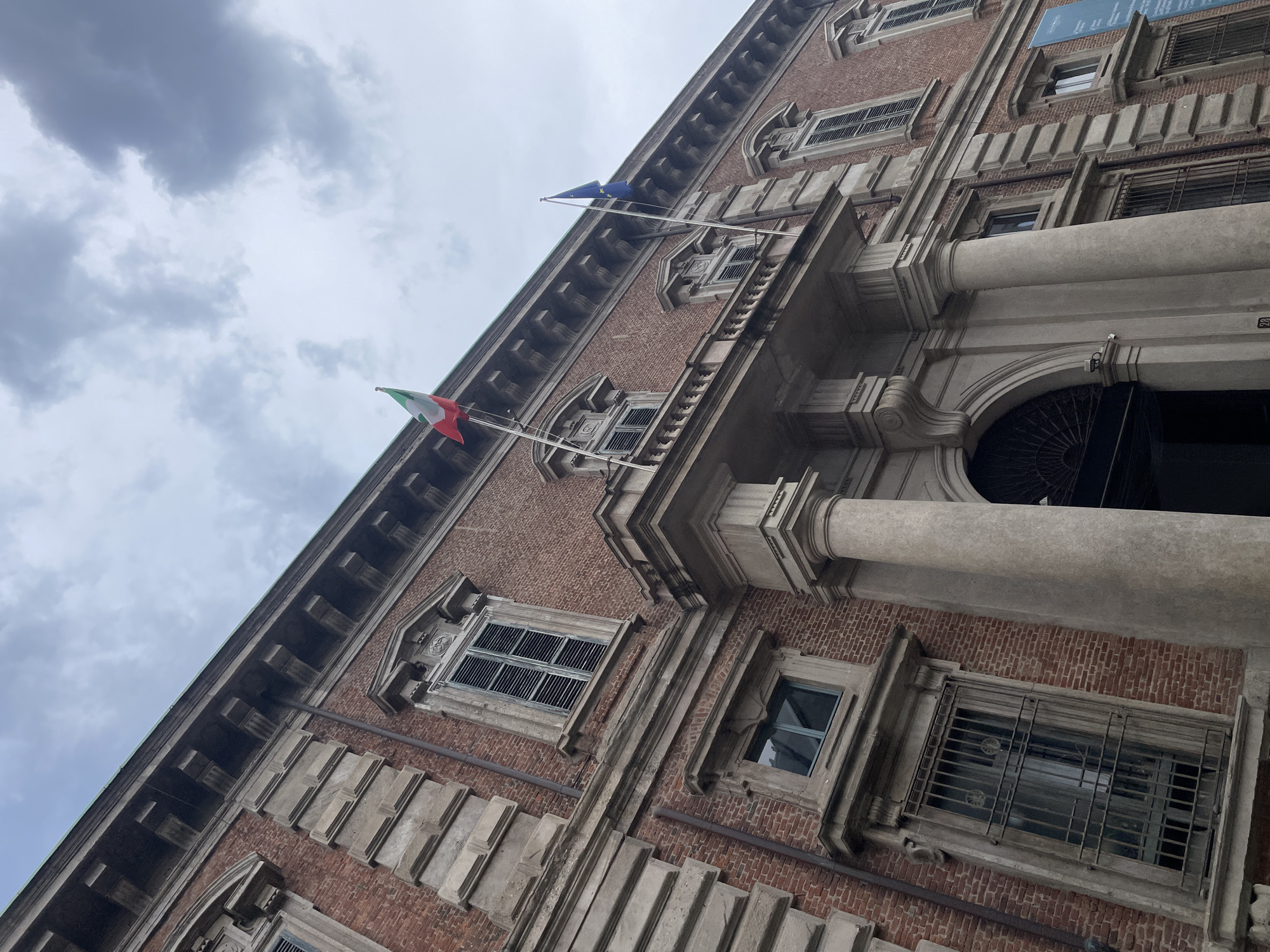 The image shows a low-angle view of the facade of Palazzo Turati in Milan, Italy. The building is constructed of red brick with gray stone architectural details. Two flags are visible: the Italian flag and the European Union flag, both mounted on poles. The sky is overcast with gray clouds. The building's entrance features large stone columns and an arched doorway. Windows are spaced regularly along the facade, some with metal bars or shutters.