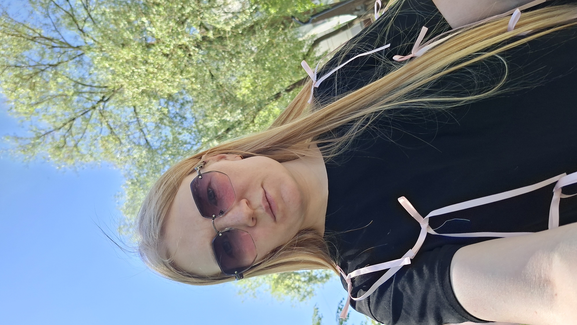 A woman is outdoors on a sunny day. She has long blonde hair, is wearing sunglasses with a pink tint, and a black shirt. Her hair and shirt are adorned with light pink ribbons. Above her, there are trees with green foliage, and a patch of blue sky. The location is the city of Chaikovsky, Russia.