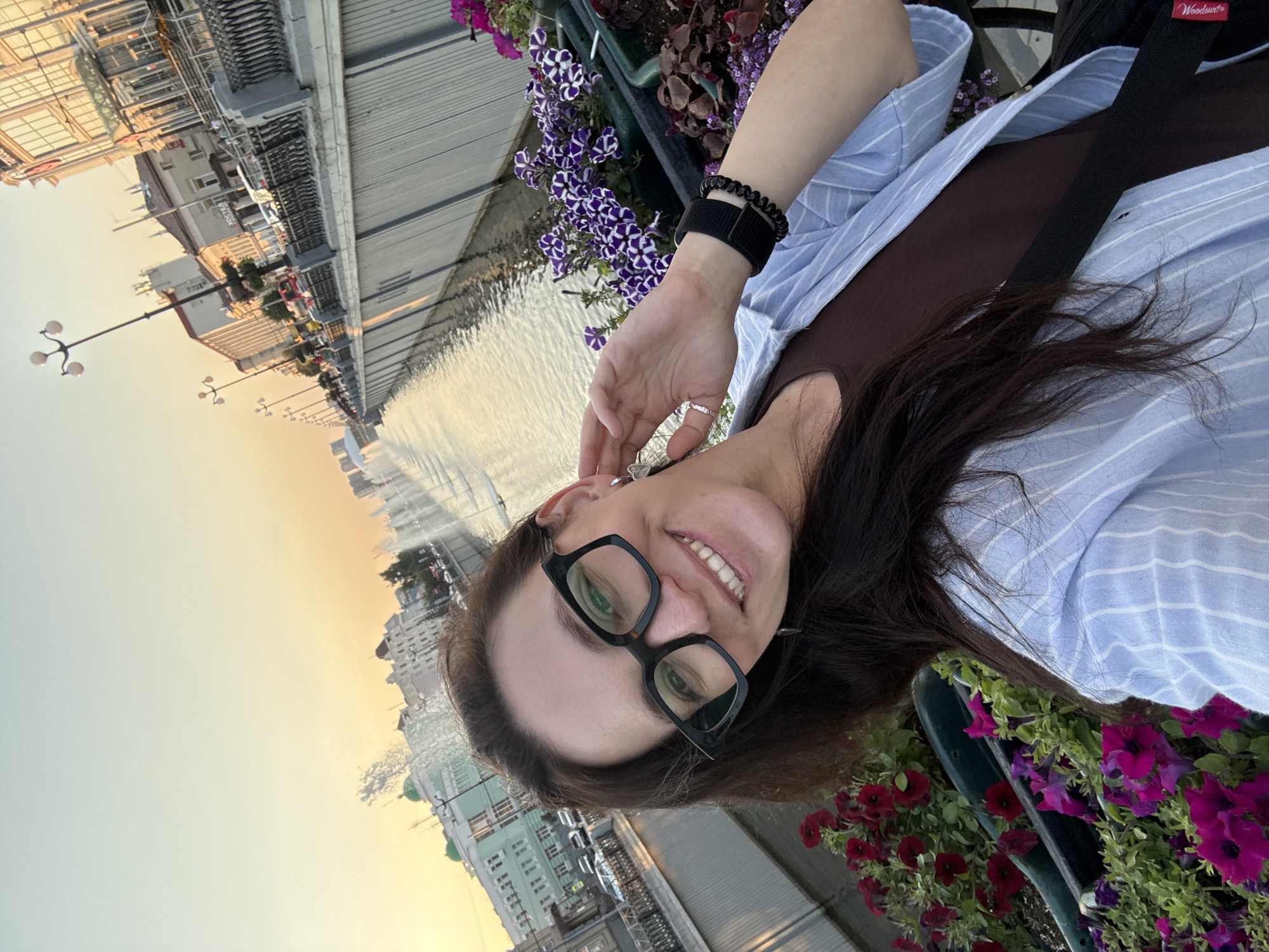 A woman with dark hair and glasses takes a selfie outdoors during sunset. She is wearing a brown tank top and a light blue striped open button-up shirt. She is smiling and touching her right ear. In the background, a canal with fountains runs through a city with buildings and streetlights. Flower planters with red and purple petunias line the canal. The sky is a gradient of pale blue and orange. The scene appears to be the Kazanka River embankment in Kazan, Russia.