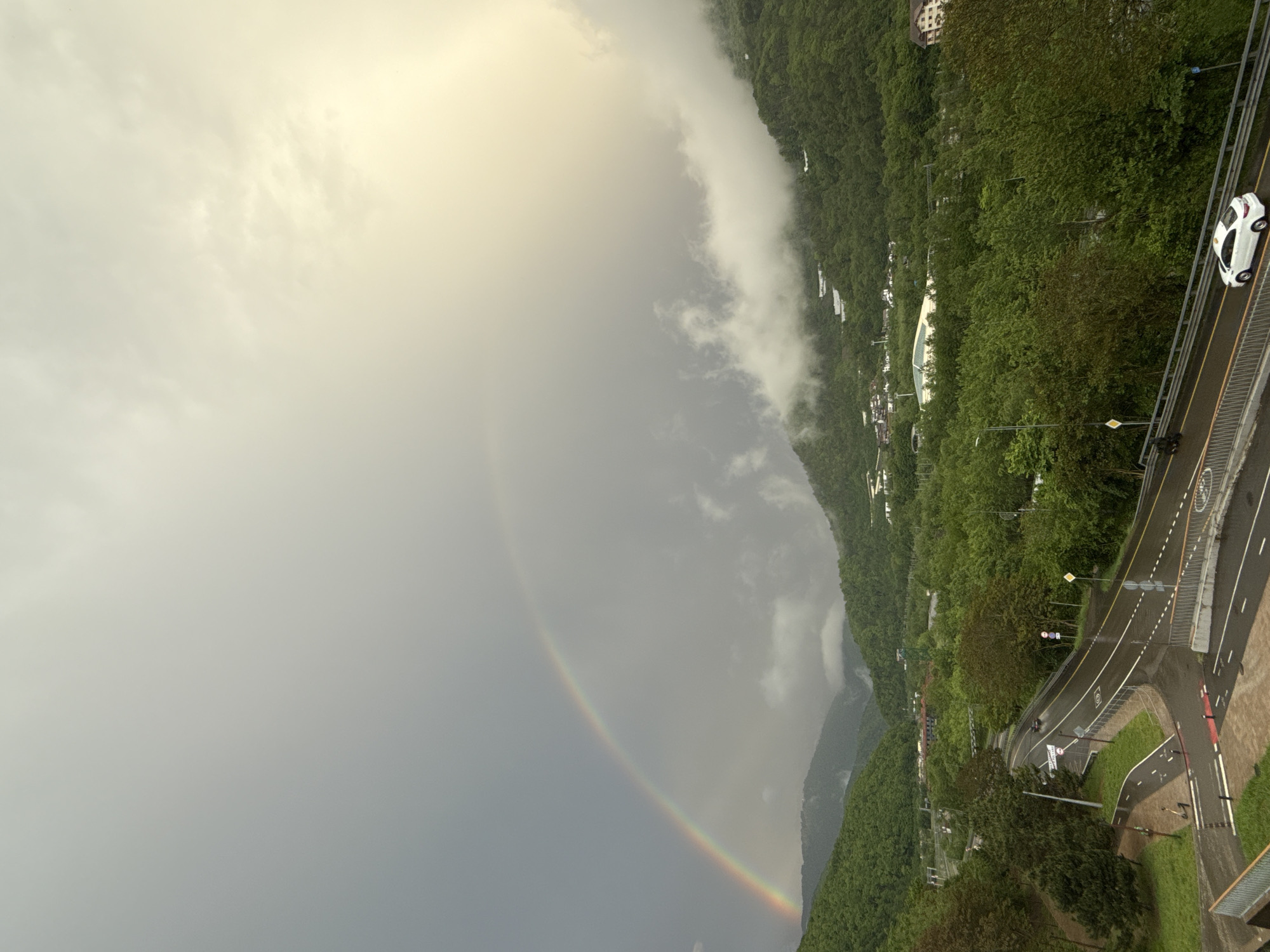 The scene depicts a partial rainbow arc in the sky over Krasnaya Polyana, Russia. The arc displays the typical colors of a rainbow. In the foreground, a road winds along a hillside covered with dense green trees. A white car is seen traveling on the road. The sky is overcast, with clouds obscuring the top of the landscape.