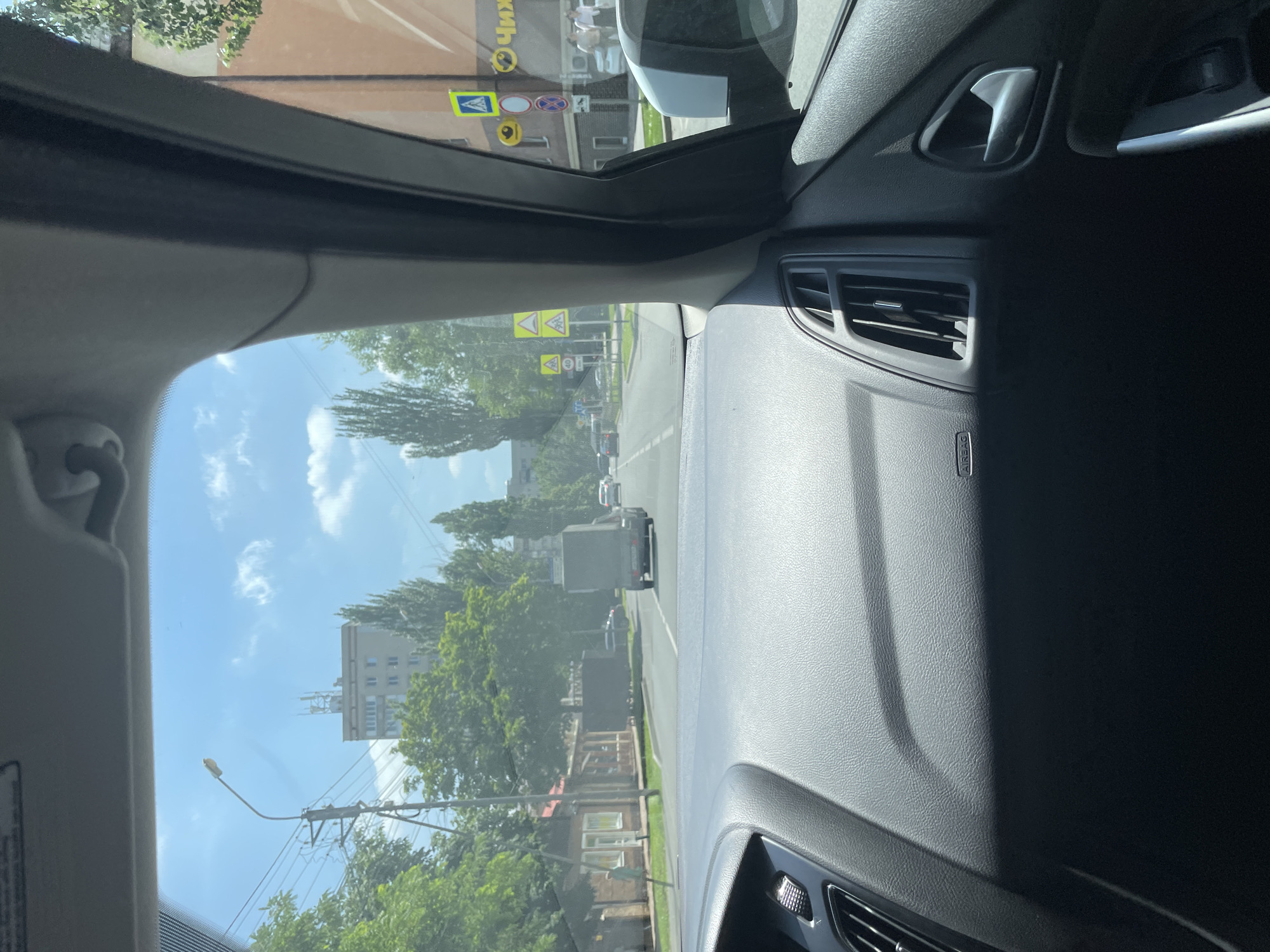 The image is taken from inside a car looking out the windshield at a street scene in Stavropol, Russia. The dashboard of the car is in the foreground, with an air vent and the "AIRBAG" label visible. The street is lined with trees and buildings, including a multi-story building in the distance. There are several cars and a truck on the road. Traffic signs are visible, including a speed limit sign indicating 40. The sky is blue with some clouds.