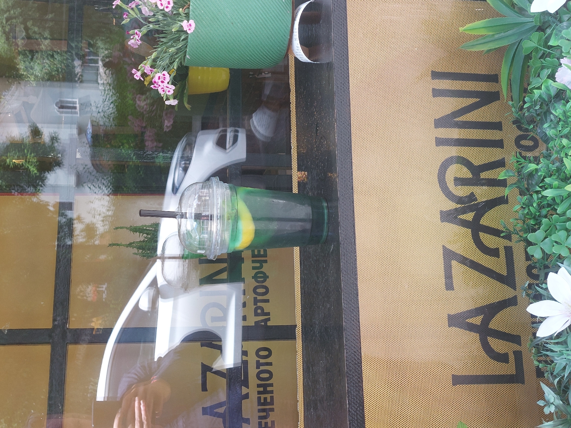 A still life composition is presented, apparently photographed through a glass surface, likely a window. On a black, framed ledge rests a green-colored drink in a clear plastic cup with a lid and black straws. The drink contains what appears to be a slice of yellow lemon.
To the left of the drink, there's a partial reflection on the glass. The reflection suggests a building across the street and a light-colored car, possibly white. The reflection also shows a person's reflection, with visible fingers.
To the right of the drink, there's a textured surface with the name "LAZARINI" printed vertically in black, next to green plants with white flowers. Below the drink, there's another name printed in Bulgarian, possibly "ЕЧЕНОТО АРТОФЧЕ".
The scene appears to be photographed from inside a cafe or restaurant. Based on the name "LAZARINI" in Bulgarian and the overall ambiance, the location is most likely in Burgas, Bulgaria, as it is a common Bulgarian surname. The environment is well-lit, suggesting daytime.