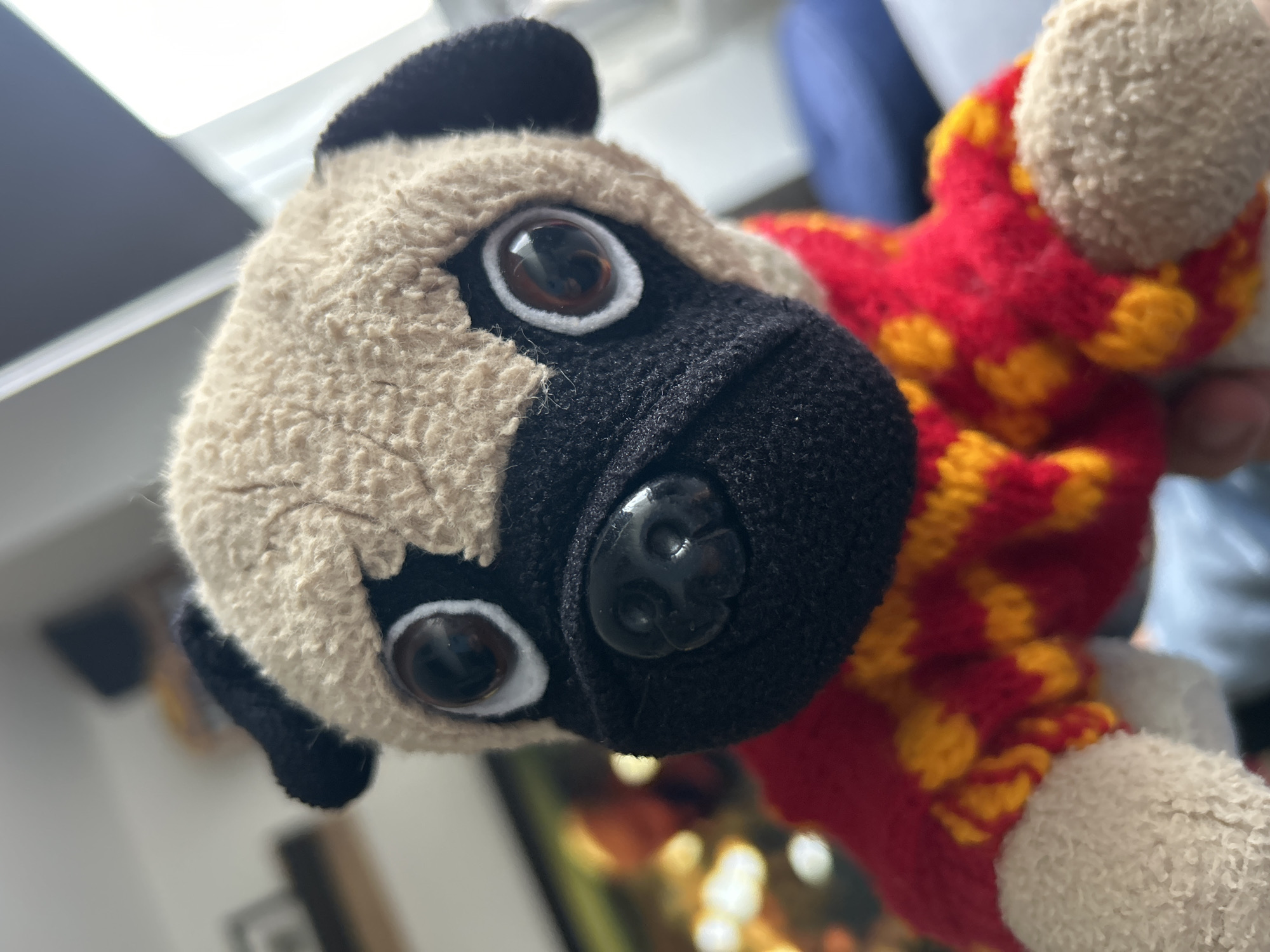A close-up image focuses on a plush toy resembling a pug dog. The pug's head is off-white with darker markings, including black ears and a muzzle. It features prominent, round, brown eyes with white rings around them. The nose is black and made of plastic with detailed features. Part of the pug's body, which is made of a red and yellow patterned knitted fabric, is also visible. The image is taken in an indoor setting, with a blurred background showing a window and other objects. The environment does not contain information specific to Huixquilucan de Degollado, Mexico.