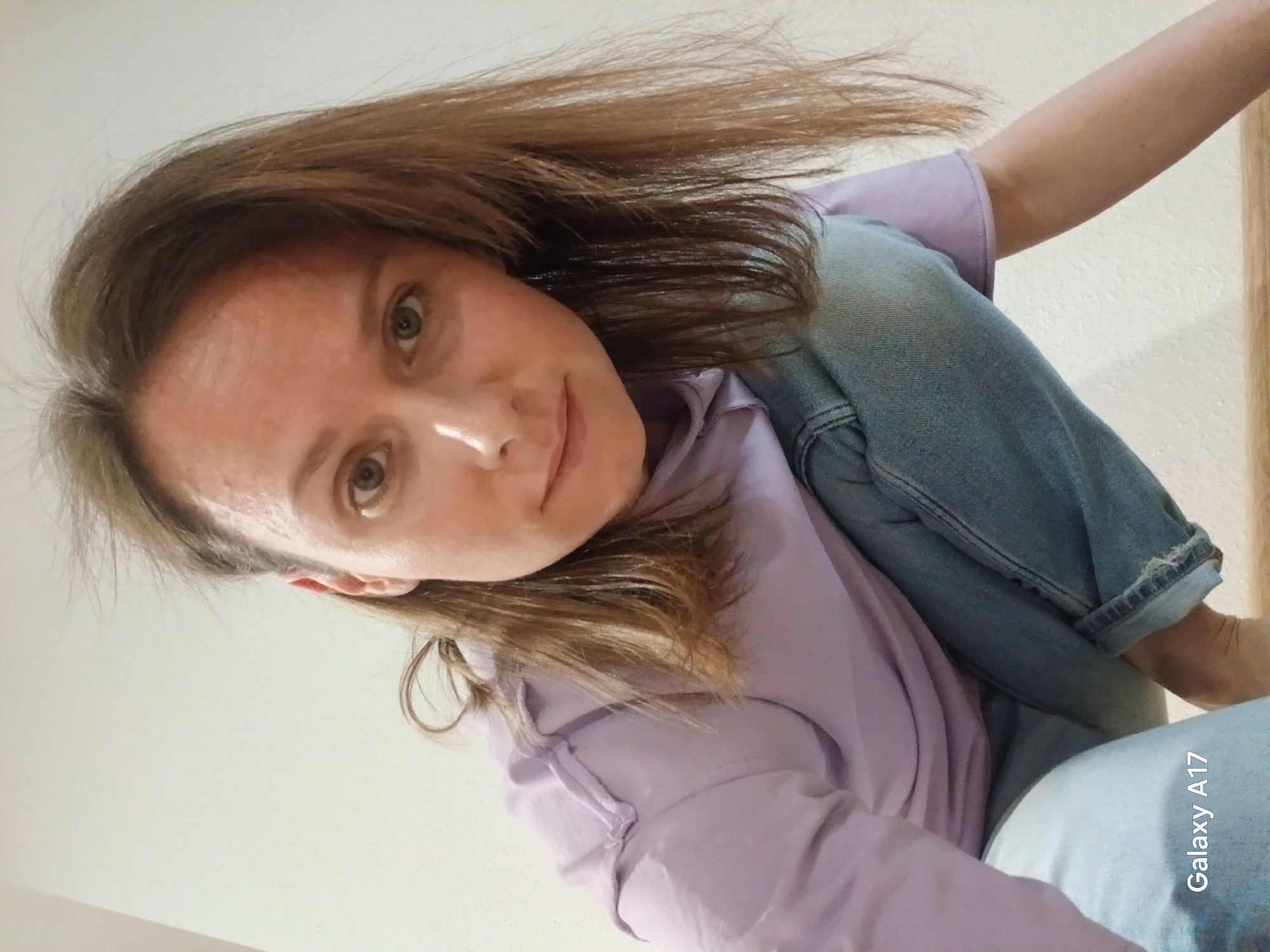 A person is pictured in an angled, close-up shot. The individual has light brown hair, fair skin, and light-colored eyes. The person is wearing a light purple top and a light blue denim jacket. The person is indoors. The location is not discernable from the image. The photo appears to have been taken with a Galaxy A17 smartphone.