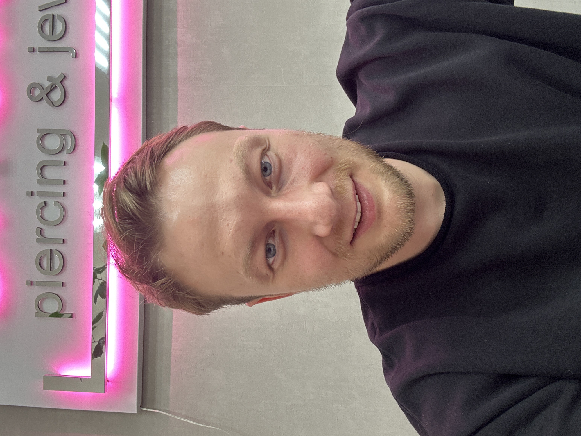 The image, originally oriented 90 degrees counter-clockwise, depicts a man taking a selfie indoors. He is a light-skinned individual with short, light brown hair and blue eyes, sporting a short beard or stubble. A friendly, gentle smile is visible on his face as he looks directly at the camera. He is dressed in a dark, likely black or deep navy, long-sleeved top or sweatshirt.
The background reveals a light-colored wall featuring prominent raised silver lettering, illuminated by vibrant pink neon lights. The visible text clearly reads "piercing & jew", indicating the establishment is a "piercing & jewelry" or "piercing & jewels" shop, located in Sochi, Russia, as per context. A vertical reflective element, possibly a mirror or polished metal, separates sections of the lettering and shows a subtle reflection of some greenery. The pervasive pink light influences the overall atmosphere and color cast on the man's hair. The scene is set indoors, with artificial lighting providing the main illumination.