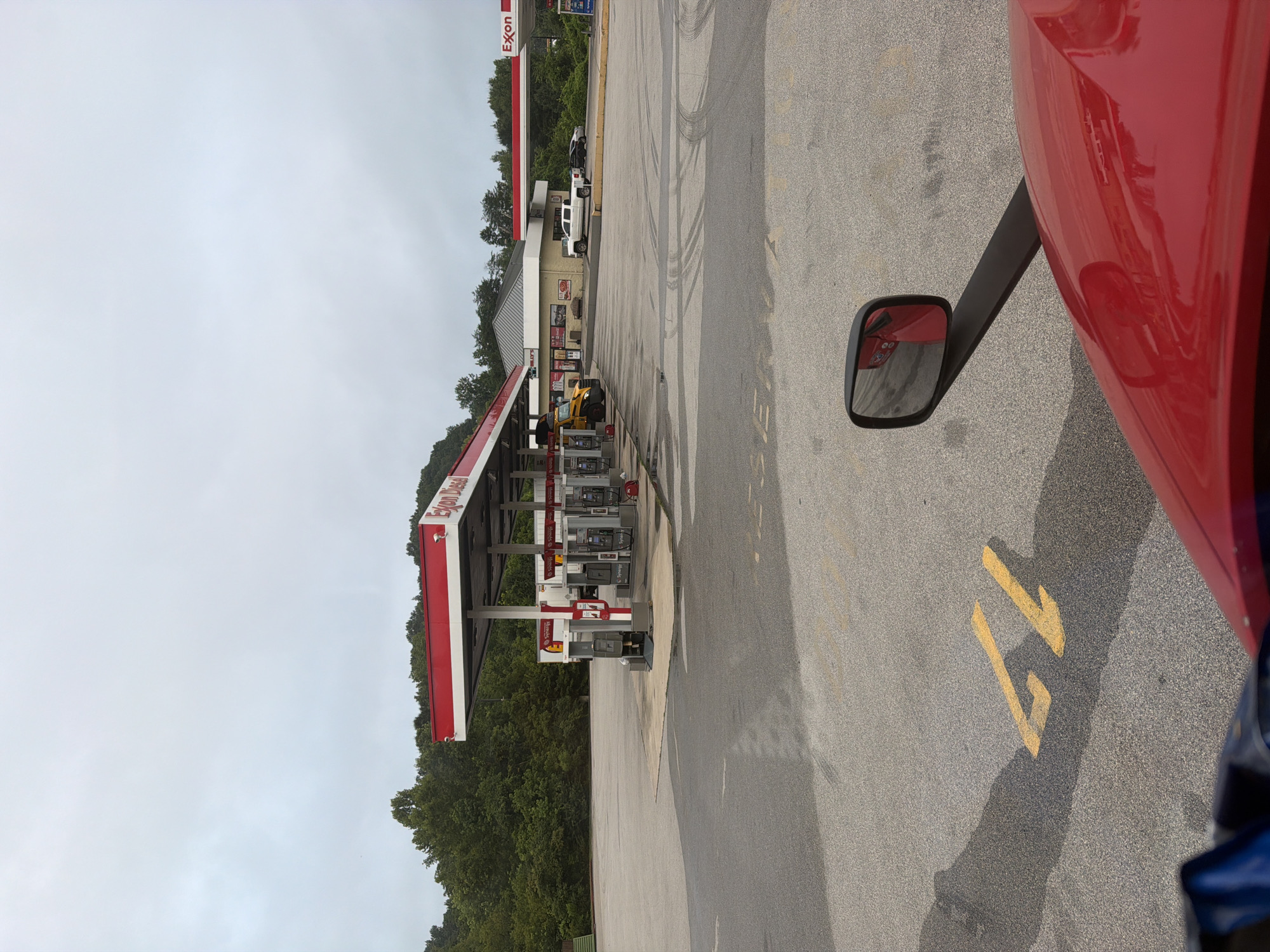 A red truck cab is in the foreground, with a side mirror reflecting part of the truck and the scene behind it. The truck is parked in an asphalt lot at an Exxon gas station in Wildwood, United States. Several gas pumps with red awnings and "Exxon Diesel" signage are visible. A yellow school bus, a white pickup truck, and two dark sedans are parked in the lot. In the background, a wooded area with green trees is present. The sky is overcast and cloudy. Yellow painted numbers "11" are visible on the asphalt in front of the truck.