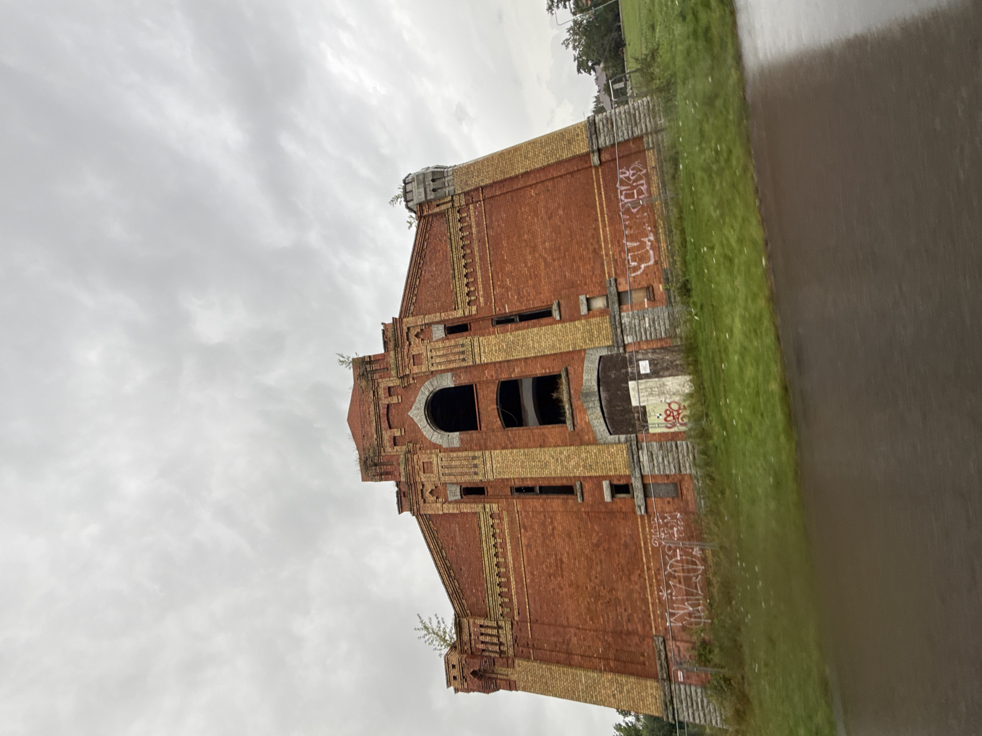 An imposing, abandoned brick building with yellow brick accents and arched windows stands under a cloudy sky. The building exhibits signs of decay, with graffiti visible on its lower walls and a chain-link fence in front. Overgrown vegetation is present on the rooftop and in the surrounding grassy area. A wet, dark surface, likely a road or pavement, occupies the foreground. The scene appears to be an industrial or historical structure, possibly a former factory or public building, in a state of disuse. The specific location within Tallinn, Estonia, cannot be determined from the image alone.