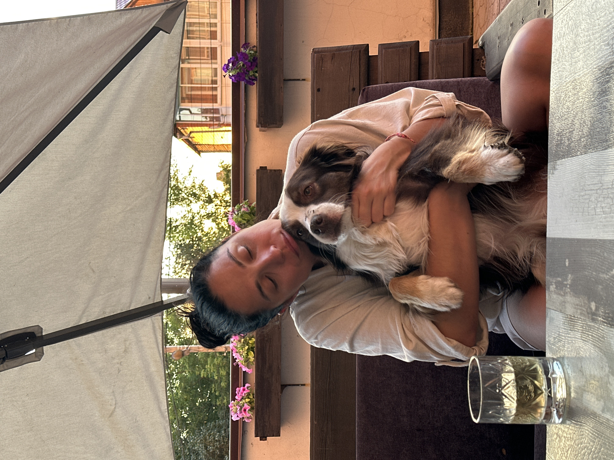 In Irkutsk, Russia, on a patio under a large umbrella, a woman with dark hair pulled into a bun holds a medium-sized brown and white dog. The woman is wearing a light-colored oversized shirt and shorts. She is leaning her head towards the dog, her eyes closed, in a gesture of affection. The dog is looking towards the camera. In the foreground, a glass of amber liquid sits on a wooden table. In the background, there are flower boxes with pink and purple flowers, wooden benches, and a building. The scene appears to be a relaxed outdoor setting.