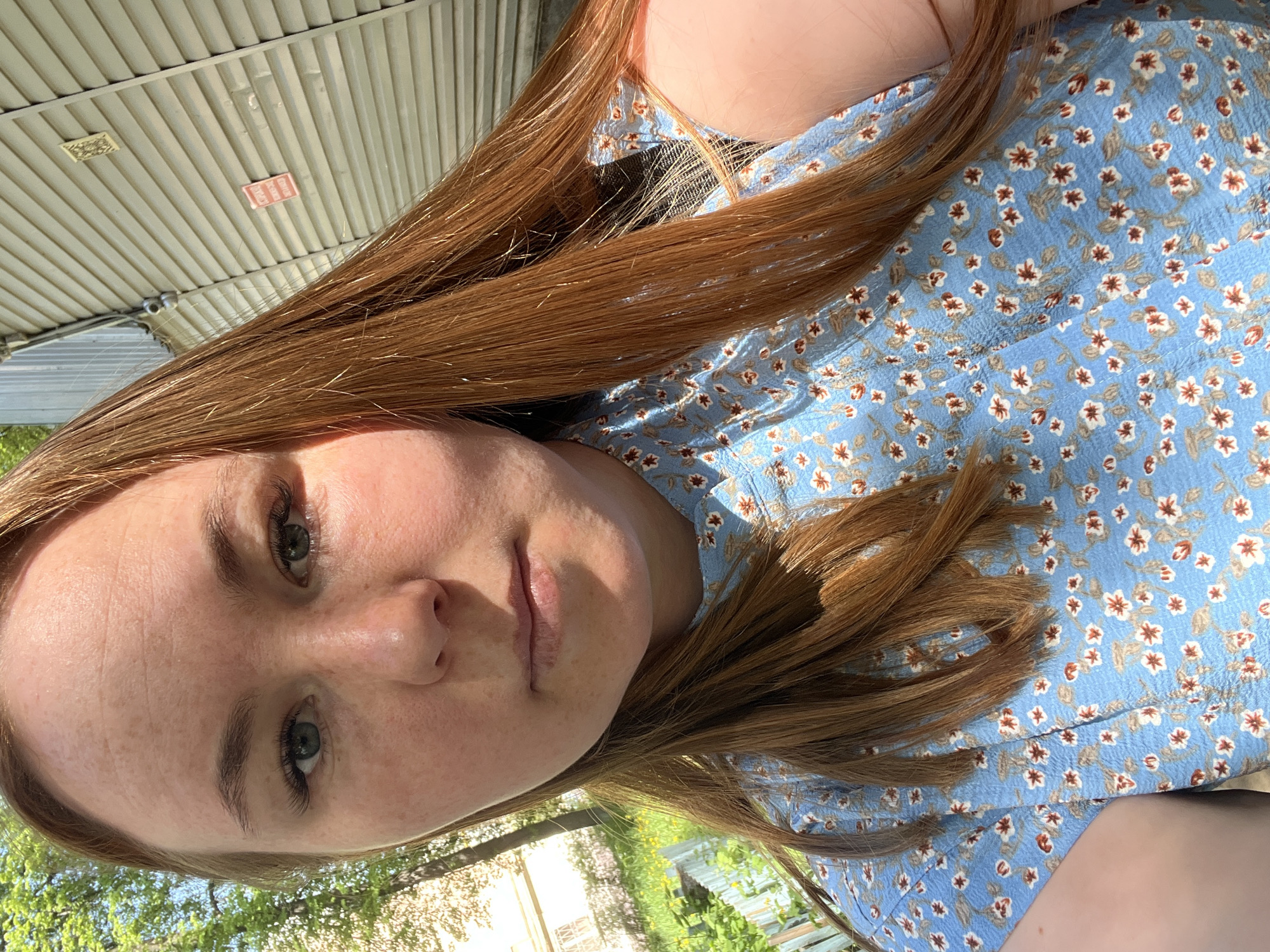 A young woman with light skin and red-brown hair is facing the camera. She has fair skin, visible freckles, and green eyes. She is wearing a blue dress with a small floral pattern of white and brown flowers.
The background shows a corrugated metal awning, suggesting an outdoor setting. Some greenery and a blurry cityscape are visible in the bottom left corner, implying an urban environment. The image appears to be taken in daylight with natural lighting. The photo appears to have been taken near Chaikovsky, Russia, since that was specified for the location.