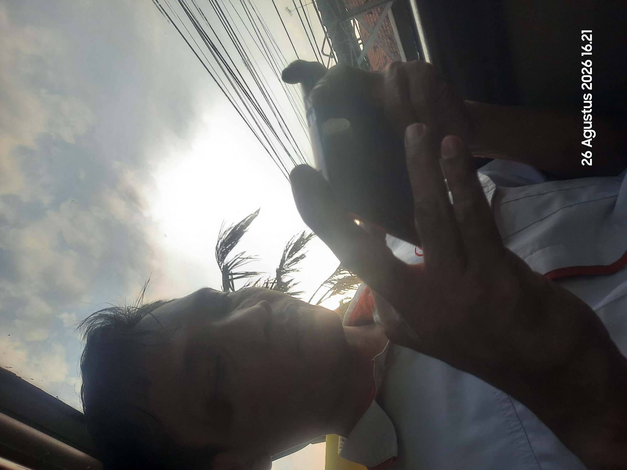 In Sukokerto, Indonesia, on August 26, 2026, at 16:21, a close-up shot captures a person's profile as they look down at their smartphone, held in their hands. The individual, with dark hair and fair skin, is wearing a white collared shirt with an orange trim visible at the collar. The lighting suggests it is late afternoon, with the sun high and partially obscured by clouds, creating a bright, hazy sky. The background shows a dense network of overhead wires, a glimpse of what appears to be a tiled roof, and palm fronds silhouetted against the sky. The focus is on the person and their device, with the surrounding environment blurred, creating a shallow depth of field.