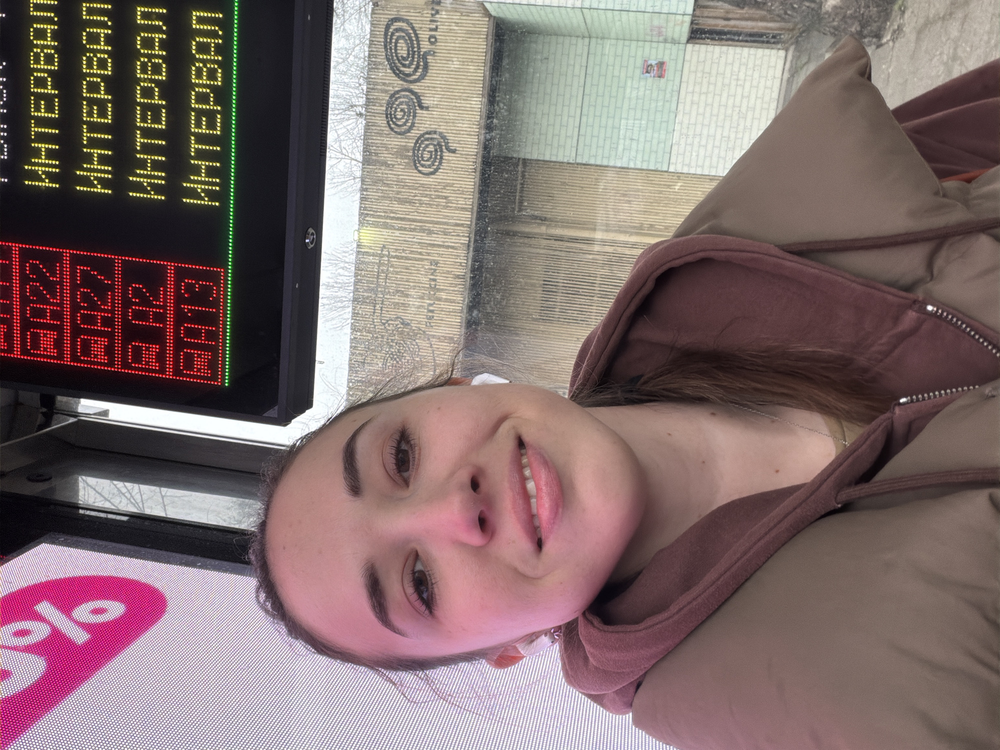 A young woman is taking a selfie on public transport in Rostov-on-Don, Russia. She is wearing a brown jacket and has an earbud in her ear. A digital display showing route information and arrival times is visible above her head. The reflection in the window behind her suggests she is near a building with graffiti.