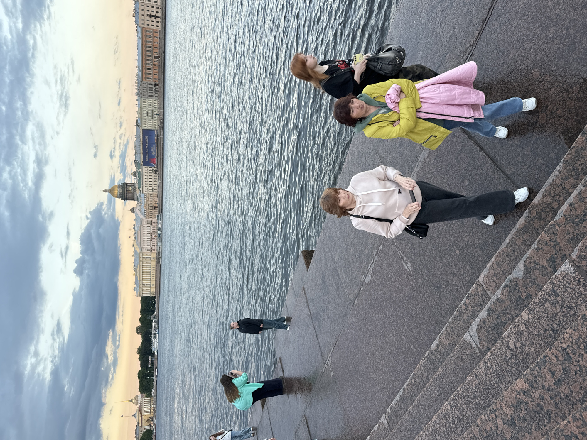 The image depicts a waterfront scene in Saint Petersburg, Russia, likely along the Neva River, with the silhouette of the city's iconic architecture in the background under a cloudy sky. In the foreground, several individuals are visible on a granite embankment. One person in a teal hoodie and dark pants stands near the water, holding a phone as if taking a picture. Another person in a black hoodie and jeans stands nearby. Further to the right, three women are walking. One woman in a light-colored hoodie and dark pants, another in a bright yellow jacket over a green hoodie with a pink garment draped over her arms, and a third woman with red hair wearing a black shirt. The water is choppy, indicating a breezy day. The architectural skyline features classical buildings, a prominent dome, and a tall spire, suggesting a view of the historic city center.