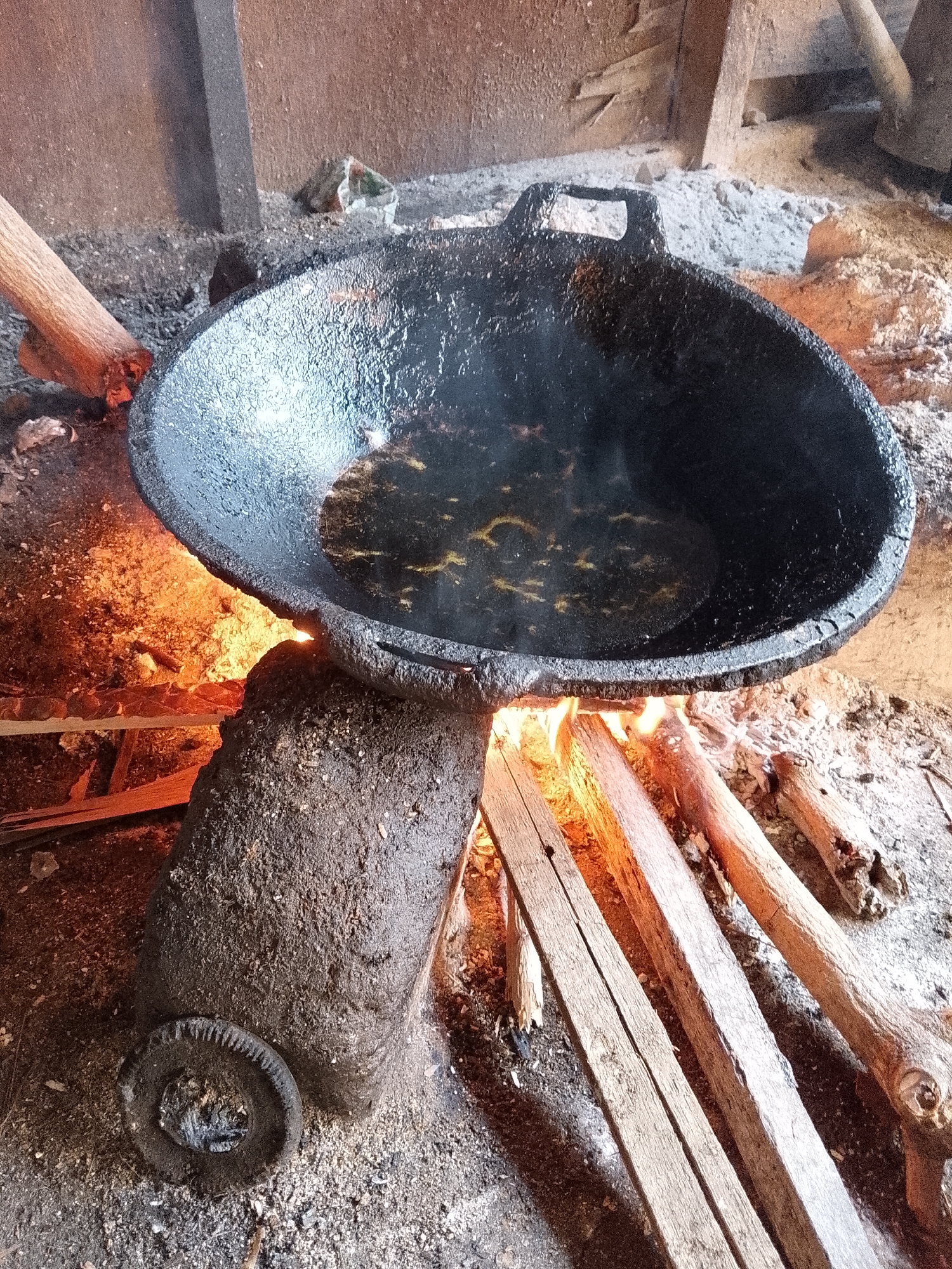 This image captures a traditional, rustic cooking setup, likely located in Lhokseumawe, Indonesia. The central focus is a large, dark, cast-iron wok or pan, which is positioned over an open wood fire. The wok contains a small amount of clear liquid, likely cooking oil, which appears to be heating, with slight reflections visible on its surface.
The fire is fueled by several wooden logs and planks laid on the ground beneath the wok, emitting bright orange flames that illuminate the surrounding area. The wok rests on a sturdy, organic-looking stove structure, seemingly constructed from hardened earth or clay, which shows signs of extensive use with its dark, textured, and somewhat soiled surface. A circular, dark object, possibly a discarded or integrated part of the stove, lies on the ground near its base in the foreground.
The ground surrounding the fire is a mixture of ashes, dirt, and small debris, indicating a well-used cooking space. In the background, hints of wooden beams or walls suggest this cooking area might be indoors or under a shelter. No people are visible, and there is no discernible text in the image. The scene conveys a sense of traditional food preparation using fundamental tools and methods.