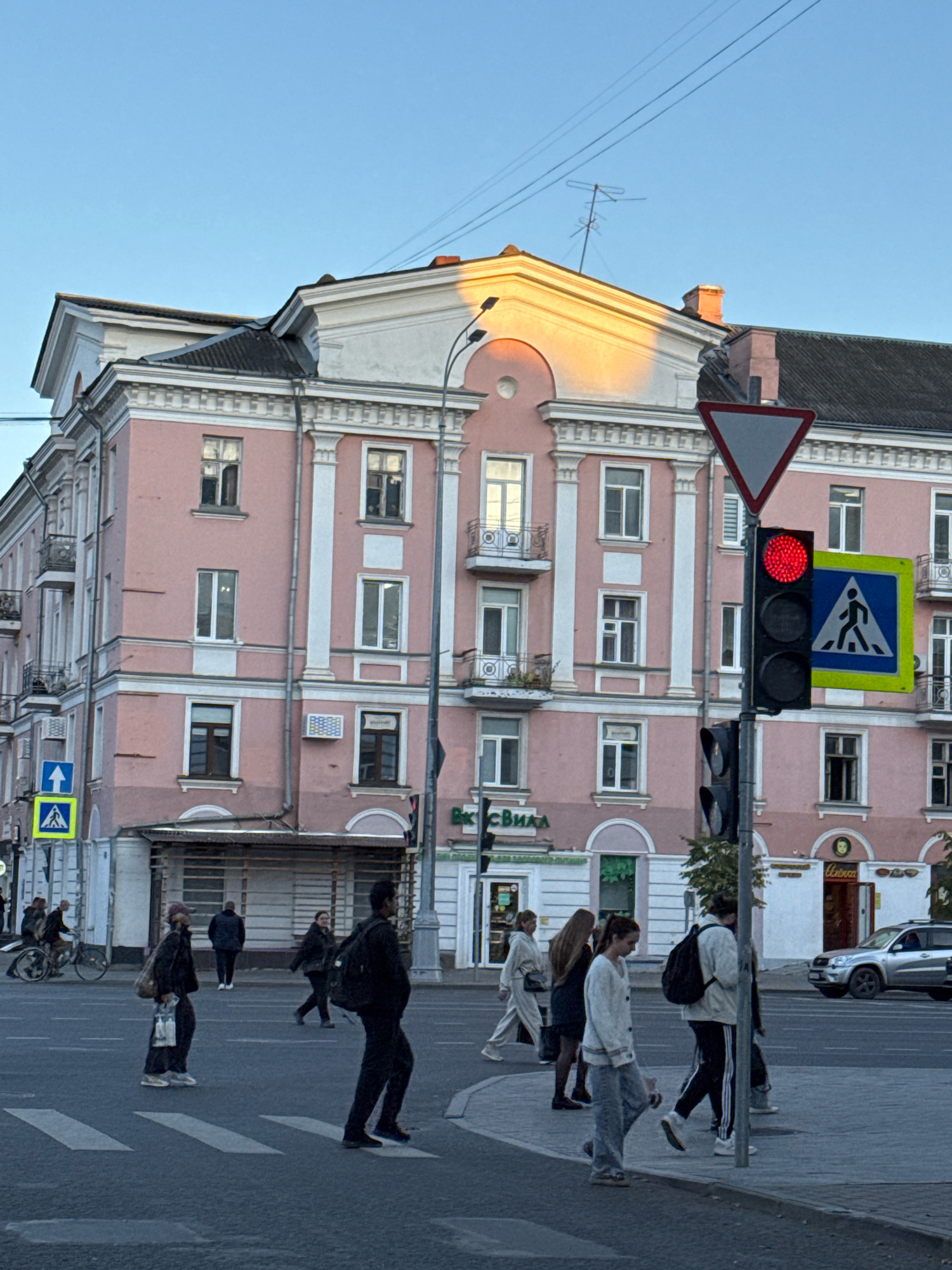The image depicts a street scene in Tver, Russia, near the intersection of Tverskaya Street and Sovetskaya Street. A large, pink apartment building with white trim dominates the background. The building features multiple stories with numerous windows, some with balconies. The sun is casting a warm glow on the upper facade of the building. A traffic light is visible, displaying a red signal, and a pedestrian crossing sign is also present. Several individuals are crossing the street at a crosswalk. On the left, a person on a bicycle is approaching the intersection. In the foreground, pedestrians are walking across the street. A silver car is parked or moving on the right side of the road. The sky is clear and blue.