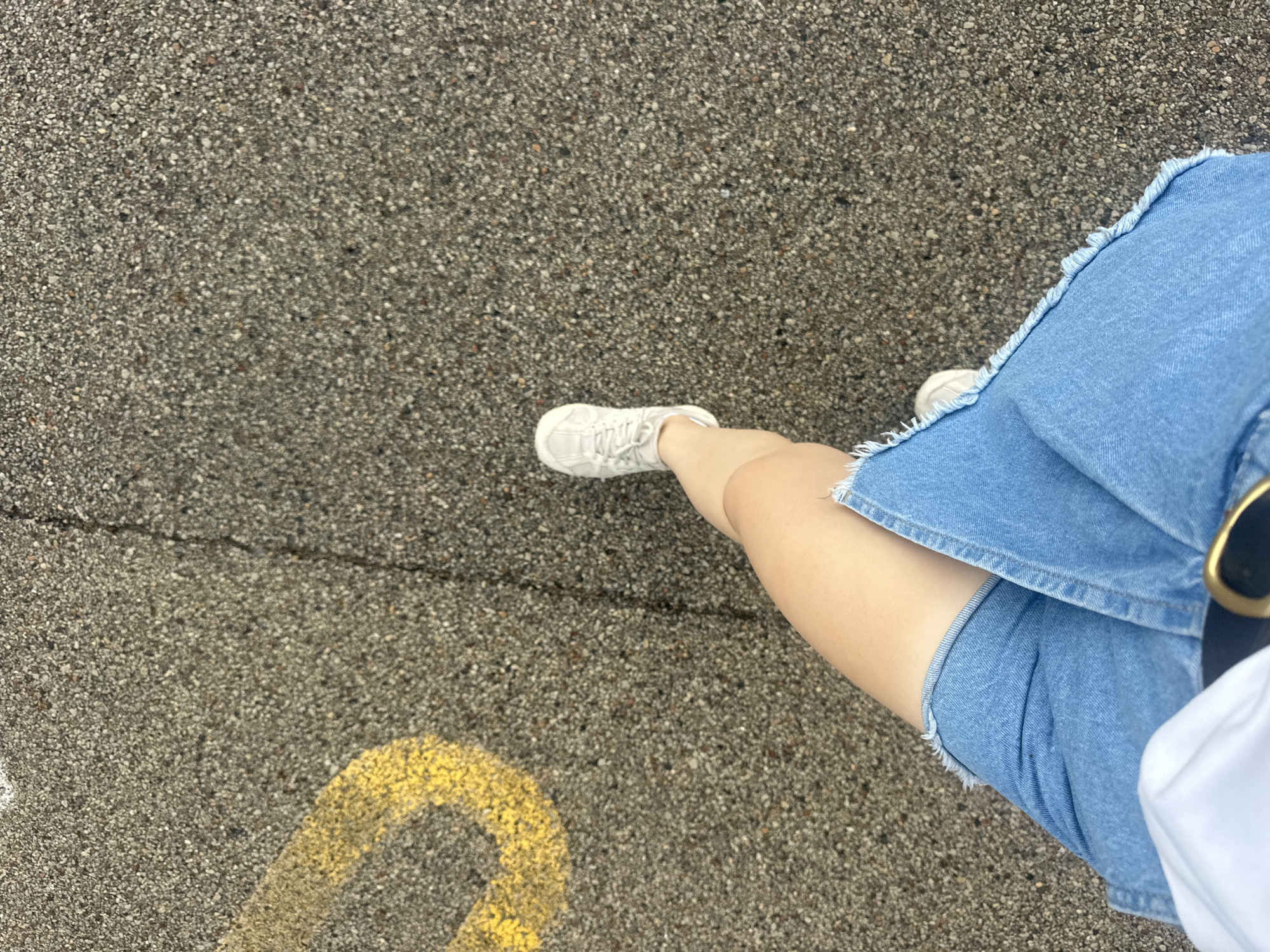 The image shows a person's legs from the thigh down, walking on an asphalt surface marked with a yellow line. The person is wearing a denim skirt with frayed edges and white sneakers. The environment appears to be outdoors, possibly a parking lot or a sidewalk in Schaumburg, United States of America, given the asphalt and markings. The lighting suggests it is daytime.