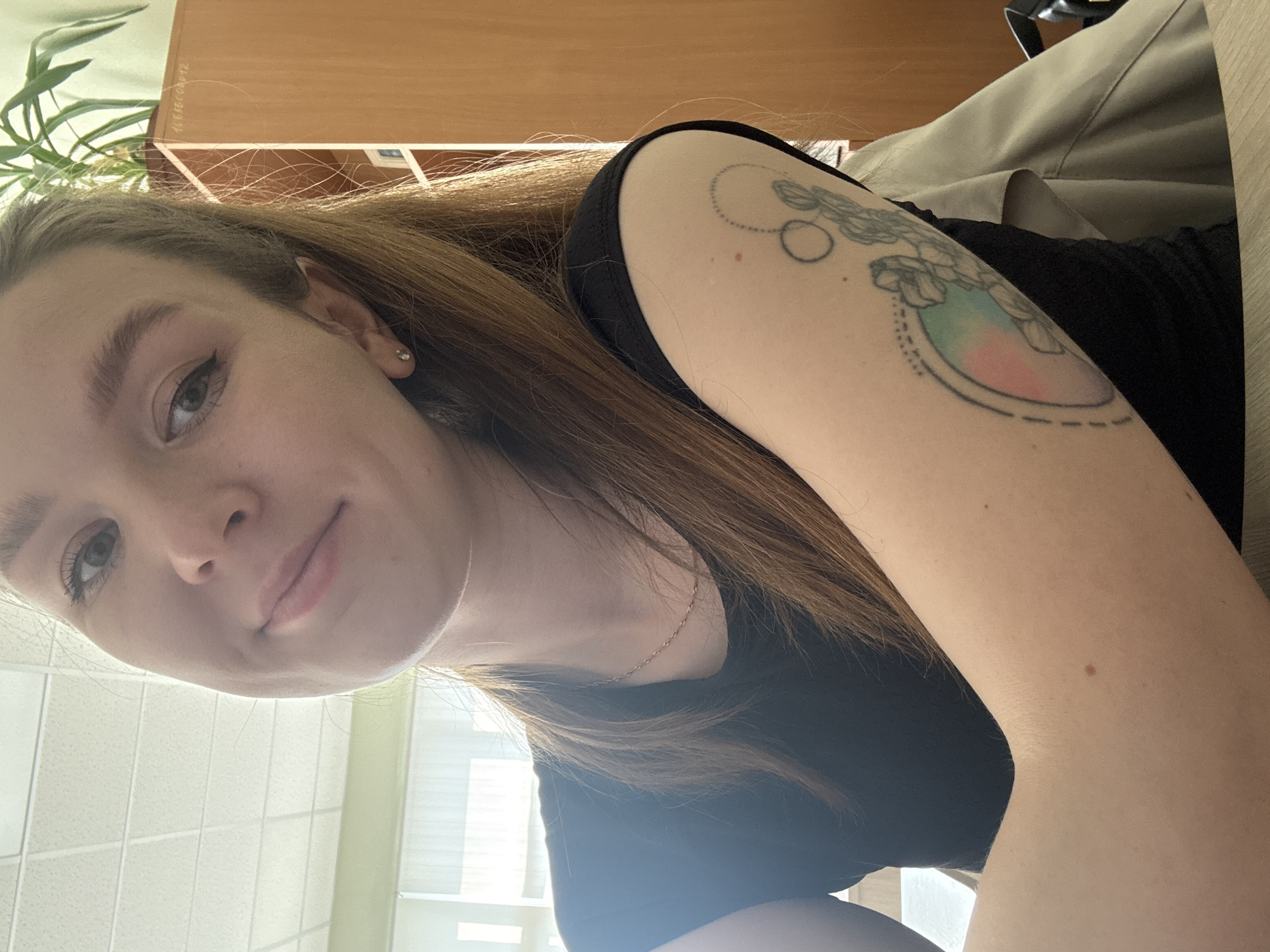 A young woman is photographed from the chest up, looking at the camera with a slight smile. She has long, brown hair and is wearing a black t-shirt. A silver necklace is visible around her neck. She has a tattoo on her left arm, which appears to be a collection of flowers within a circular frame with a gradient of pastel colors. The background shows a classroom or office environment with a white ceiling with fluorescent lighting, a wooden cabinet with some papers and a plant on top. The woman is likely in Izhevsk, Russia, as indicated by the provided information.