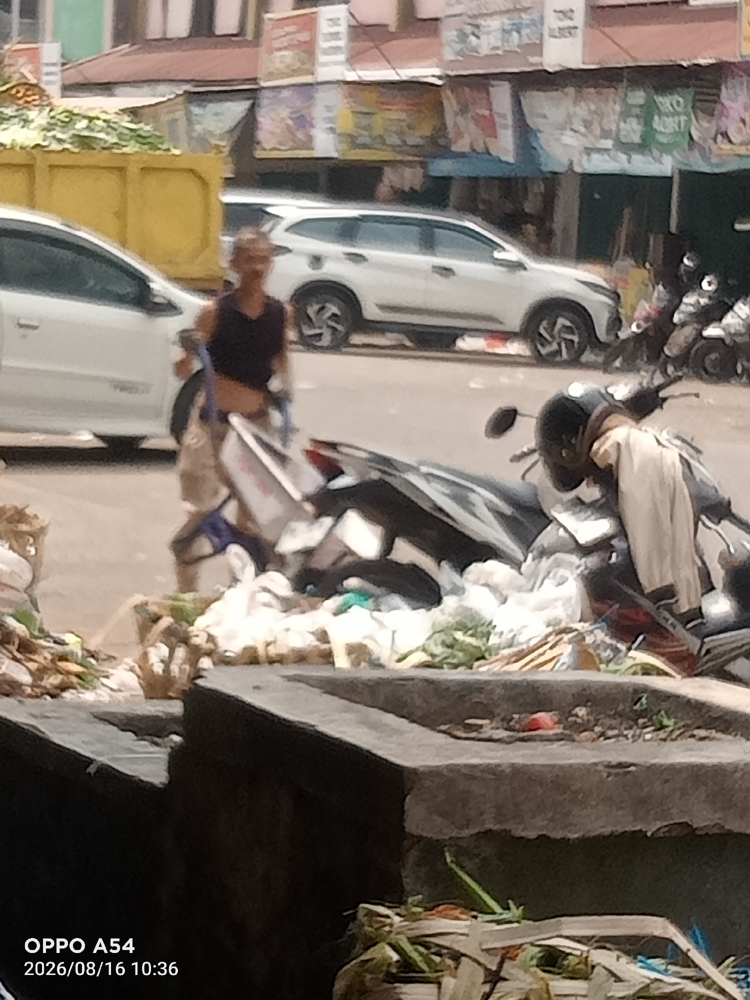 The image captures a street scene in Jambi, Indonesia. In the foreground, a person is visible, walking while carrying bags. Behind them, several motorcycles are parked, some covered with clothing. A white car is also present on the street. The background shows buildings and awnings, suggesting a commercial or residential area. The overall impression is that of an ordinary day in an urban environment, possibly a market area given the presence of the motorcycles and pedestrian activity.