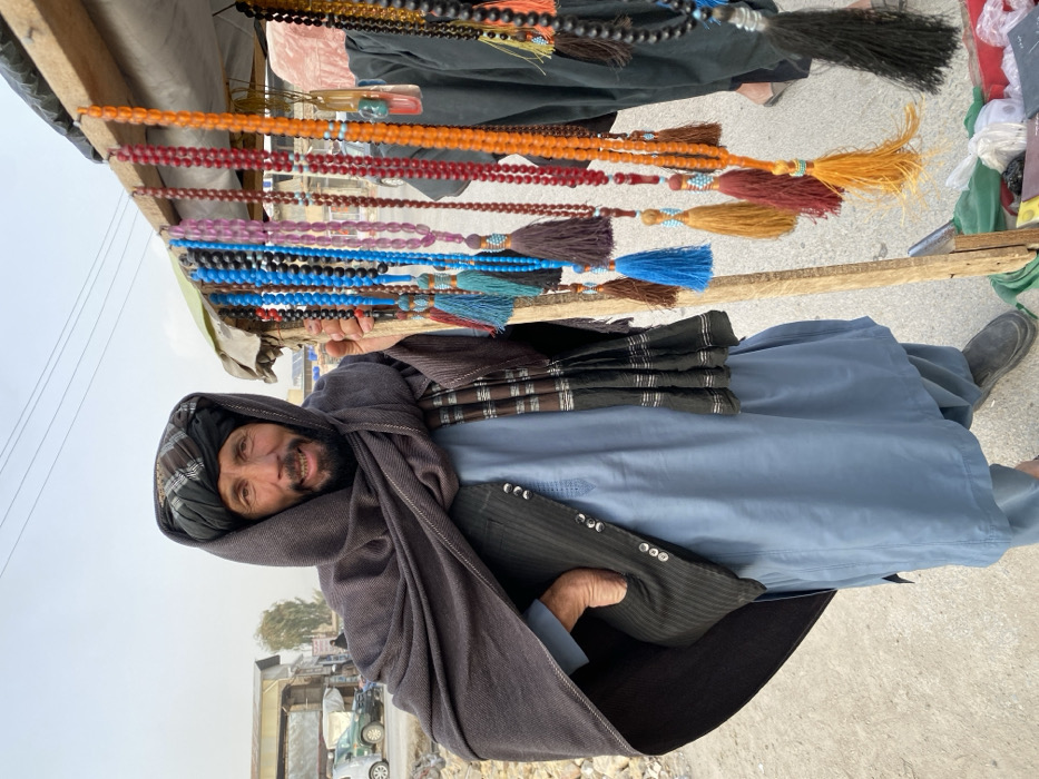 An outdoor scene in Kandahar, Afghanistan, features a man standing on an unpaved street next to a makeshift vendor stall. The man, with a dark beard and mustache, smiles toward the viewer. He wears a dark turban, a dark brown shawl draped over his head and shoulders, a light blue traditional tunic (shalwar kameez), and a dark, vertically striped vest with silver buttons. His left hand rests in his vest pocket.
To his right, a wooden stand displays numerous strands of prayer beads (tasbih/misbaha) in various colors including black, blue, pink, red, orange, and amber. Many strands feature colorful tassels in shades of blue, brown, yellow, and black, often with small light blue beaded accents. Several larger decorative pendants, including a translucent reddish-orange piece with green and blue elements, are also visible. The man's right hand rests on a wooden pole of this display.
The background shows a dusty, light-colored ground. Low-rise buildings with corrugated roofs and a light-colored SUV are visible to the left. Overhead wires are present against a hazy, pale sky. The lower body of a second individual, dressed in dark clothing, is partially visible behind the bead stand on the right.