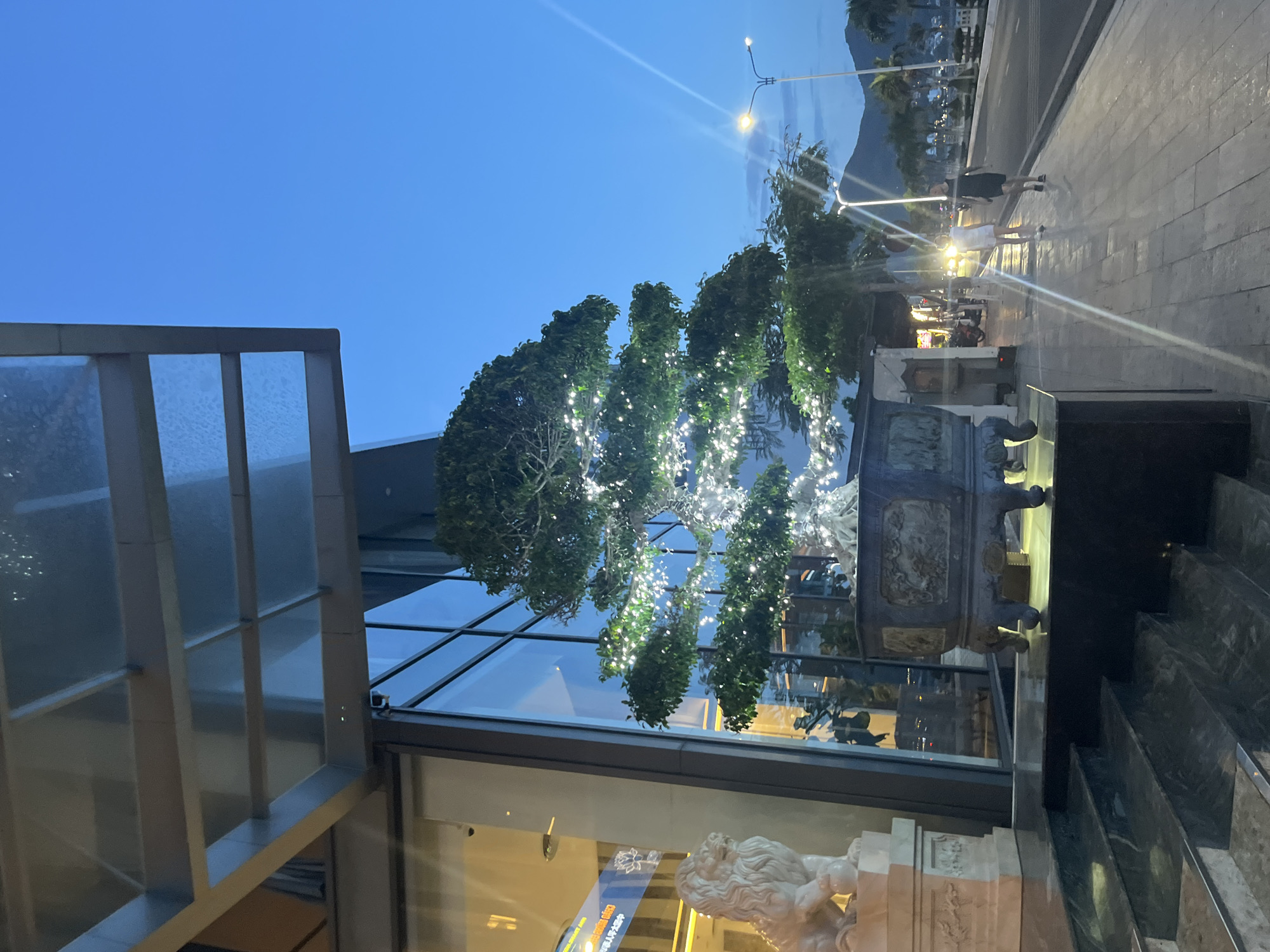 The image captures an outdoor scene in Bac Nha Trang Ward, Vietnam, at dusk or early evening. The sky is a deep blue, transitioning to darker hues.
To the left is a portion of a building's glass and metal architectural structure, reflecting the surrounding environment. Inside the building, an ornate white sculpture of a cherubic figure is visible.
In the center foreground are meticulously decorated trees illuminated with numerous small white lights, possibly for an event or festive occasion. Beside these trees is a large, dark stone pot or planter with carved detailing.
The scene extends to a street area where two people are walking on a paved walkway, with streetlights illuminating the path. Further down the street, more faint lights suggest the presence of buildings or other structures. A mountain is also faintly visible in the distance.
To the right are dark-colored steps leading up to the building, adding to the foreground's depth.
