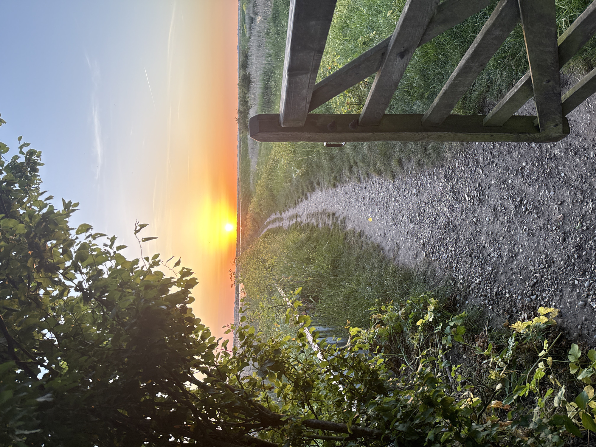 The image depicts a rural scene at sunset. A wooden gate frames a gravel path leading towards the setting sun. Vegetation lines the path, with bushes in the foreground. The sky transitions from blue to orange near the horizon. The location, Great Yarmouth, United Kingdom, is not evident from the image.