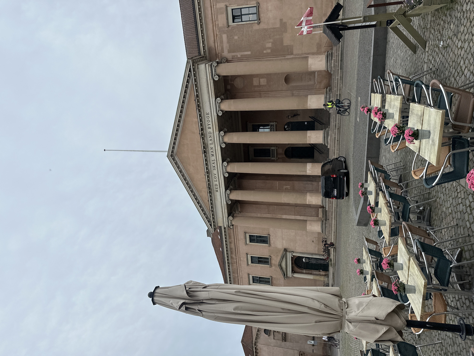 This is a photograph taken from a street-level perspective in Copenhagen, Denmark. The scene features an outdoor seating area of a cafe or restaurant in front of a large, classical-style building with a pink facade. Several tables with chairs are arranged on a cobblestone ground. Many of the tables have small potted pink flowers. In the foreground, a large, closed umbrella stands tall, taking up a significant portion of the left side of the image.
The building behind the cafe area is imposing, with a pedimented portico supported by large columns. Above the columns, text is visible on a frieze, reading "MED LOV SKAL MAN LAND BYGGE." Two Danish flags are flying from a pole on the right side of the frame. A black van is parked on the street in front of the building, and a person wearing a high-visibility vest is riding a bicycle past the building's entrance. The sky is overcast, suggesting a cloudy day. The overall impression is one of an urban public space, possibly a square or plaza, in front of an institutional or governmental building.
