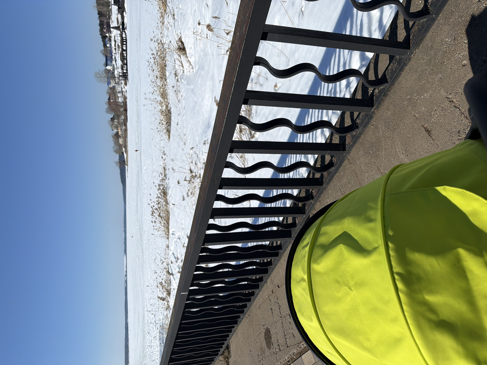 The image presents an outdoor scene overlooking the Votkinsk pond/reservoir in Votkinsk, Russia, under a clear, bright blue sky. In the foreground, the upper portion of a neon yellow stroller canopy is visible, positioned on a paved walkway. A black metal railing with a horizontal top bar and multiple wavy vertical balusters runs across the mid-foreground, separating the walkway from the frozen body of water.
Beyond the railing, the Votkinsk pond/reservoir is covered entirely in snow and ice, with patches of dry, light-brown reeds and grasses emerging from the snow. In the mid-distance on the frozen surface, a single individual, dressed in red clothing, is observed standing. The background reveals the far shoreline, featuring a scattering of residential structures, some with blue roofs, interspersed with bare deciduous trees and denser evergreen forest. A dark, wooden pier-like structure extends from the distant shore onto the frozen water. Strong sunlight illuminates the scene, casting distinct shadows from the railing onto the snow and pavement.