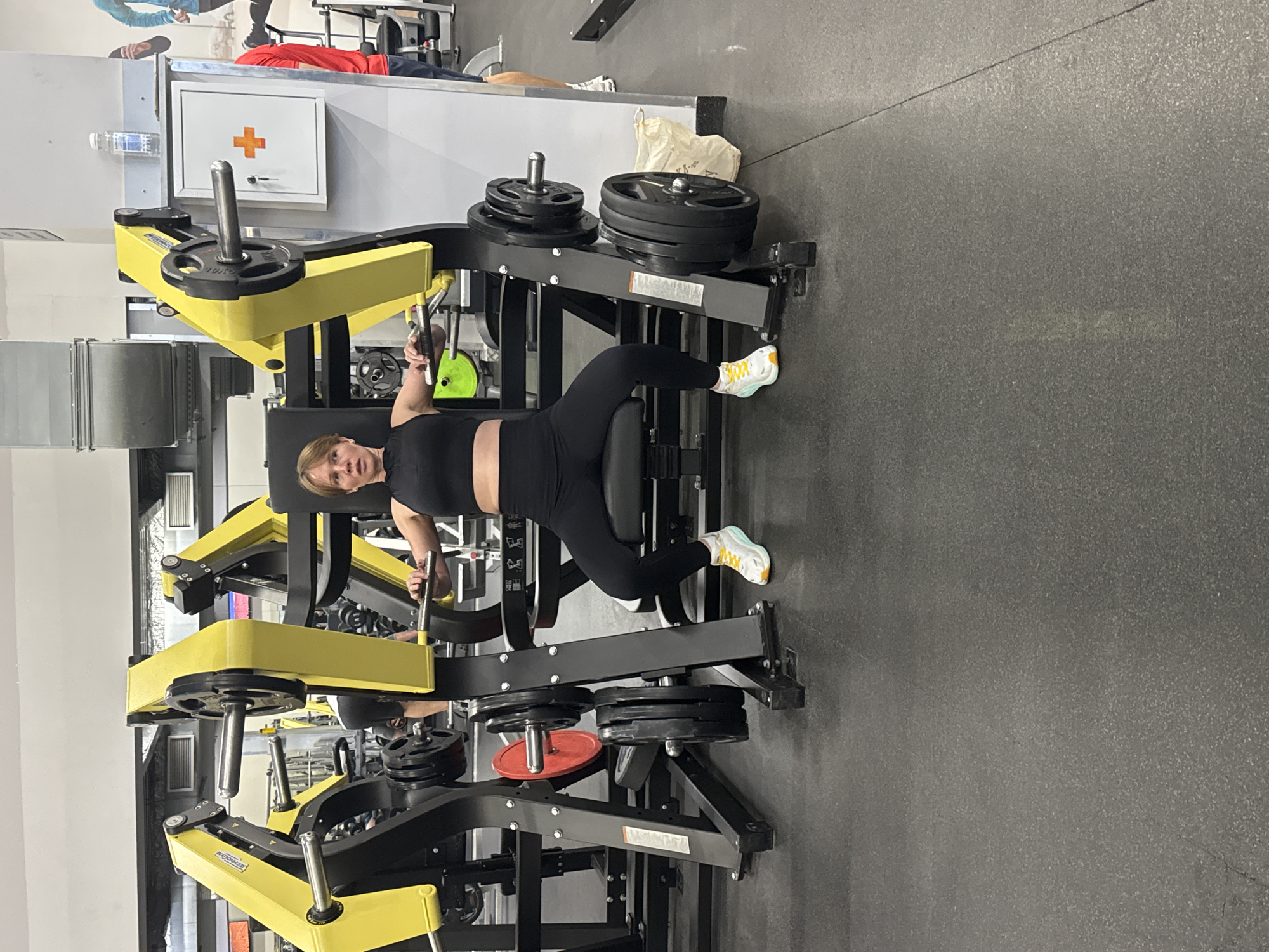A woman is positioned in a prone position on a weight machine in what appears to be a gym. The machine has yellow and black accents and is loaded with weight plates. The woman is wearing black athletic clothing and yellow and white sneakers. The gym has a gray floor and, in the background, other gym equipment and a wall-mounted first aid kit are visible. The location of Nizhny Novgorod, Russia is not ascertainable.