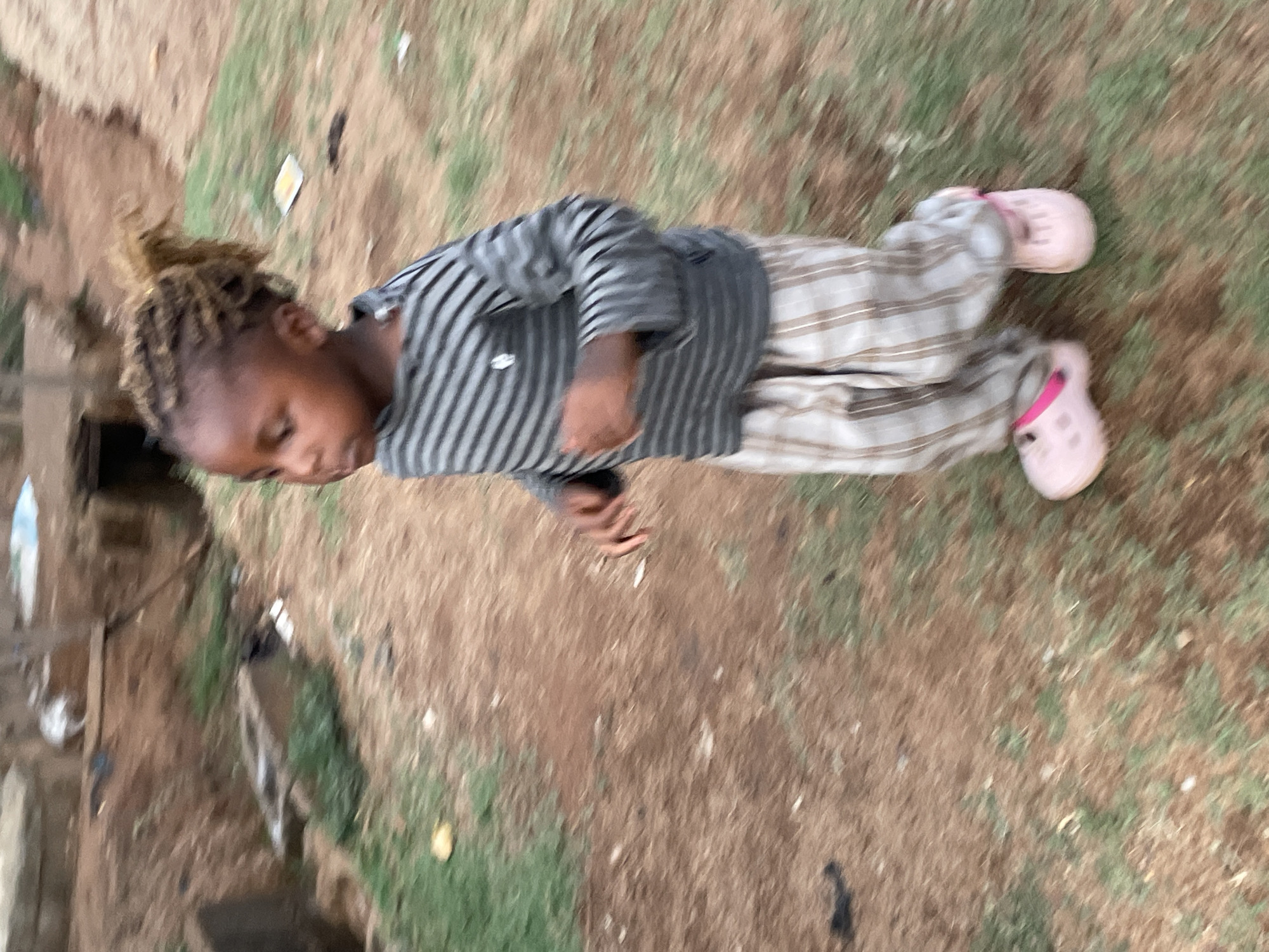 A child is lying on the ground in Barakin Akawo, Nigeria. The ground appears to be a mix of dirt and sparse grass. The child is wearing a striped shirt, striped pants, and pink Crocs. The child is facing to the left of the frame, with their head near the top. In the background, blurred structures suggest a residential or populated area.