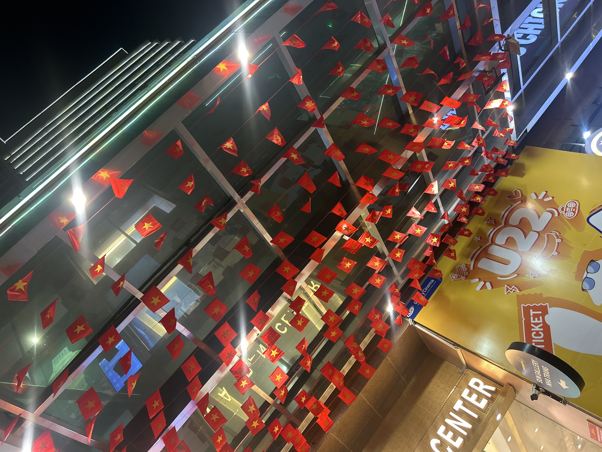 The image captures a night scene featuring the illuminated facade of a modern building, likely a shopping mall or entertainment complex, adorned with numerous small Vietnamese flags. Hundreds of red flags with yellow stars are strung in parallel lines, creating a dense canopy that reflects light and adds a festive atmosphere. These flags are the national flag of Vietnam, confirming the location context of Nha Trang, Vietnam.
The upper left portion of the image shows bright, linear artificial lights, possibly part of the building's exterior lighting or ceiling. The glass facade of the building reflects various lights and indistinct architectural elements.
On the right side, a large, vibrant yellow advertisement dominates, featuring a cartoon character wearing sunglasses and prominent text including "U22" and "TICKET." Below this advertisement, a round, black sign with white text reads "SUN GALLERY NHA TRANG" and includes an arrow, suggesting a directional indicator within the complex. Partially visible beneath the flags and above the "Sun Gallery" sign is a blue box sign with "y Cinema" (likely part of Lotte Cinema, given its distinct logo) and "ONG | SONG Nu Tan," indicating a cinema is present. Closer to the bottom, glowing white letters spell out "CENTER," suggesting an entrance or a central hub of the establishment.
The overall scene depicts a decorated, lively commercial or entertainment venue at night, celebrating or prepared for an event, with visible text guiding visitors and promoting activities.