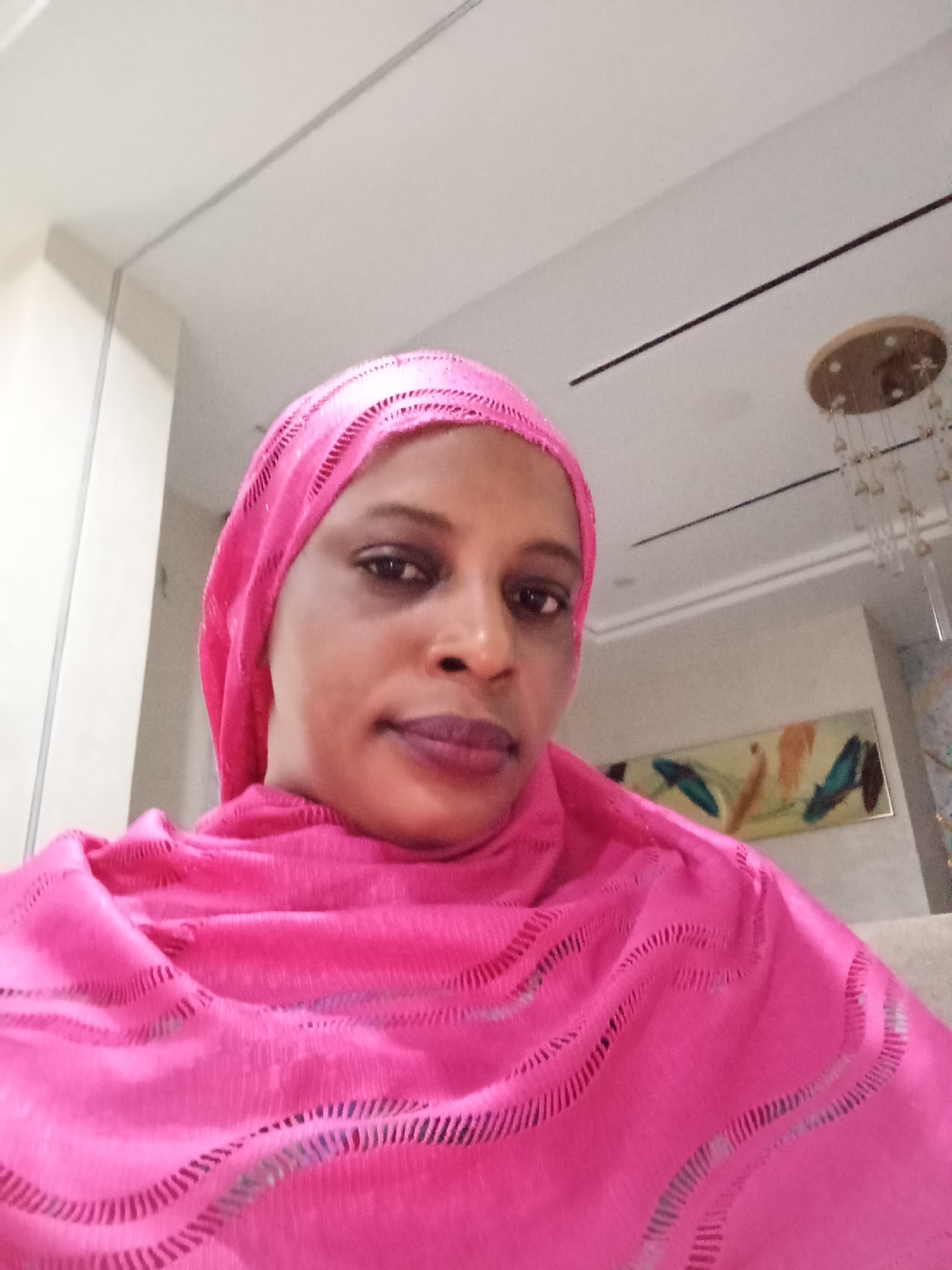 A medium shot shows a woman wearing a bright pink hijab and matching fabric draped over her shoulders. The woman's head is slightly tilted, and she is looking directly at the camera. Her lips are painted with a dark, matte lipstick. The background appears to be an indoor setting with a white ceiling, a light-colored wall, and a section of a decorative painting on the wall. A chandelier is visible hanging from the ceiling. The lighting is soft and diffused, suggesting an indoor environment. The overall scene is a portrait of the woman, likely taken in a residential or hospitality setting within Unguwar Rimi, Nigeria.