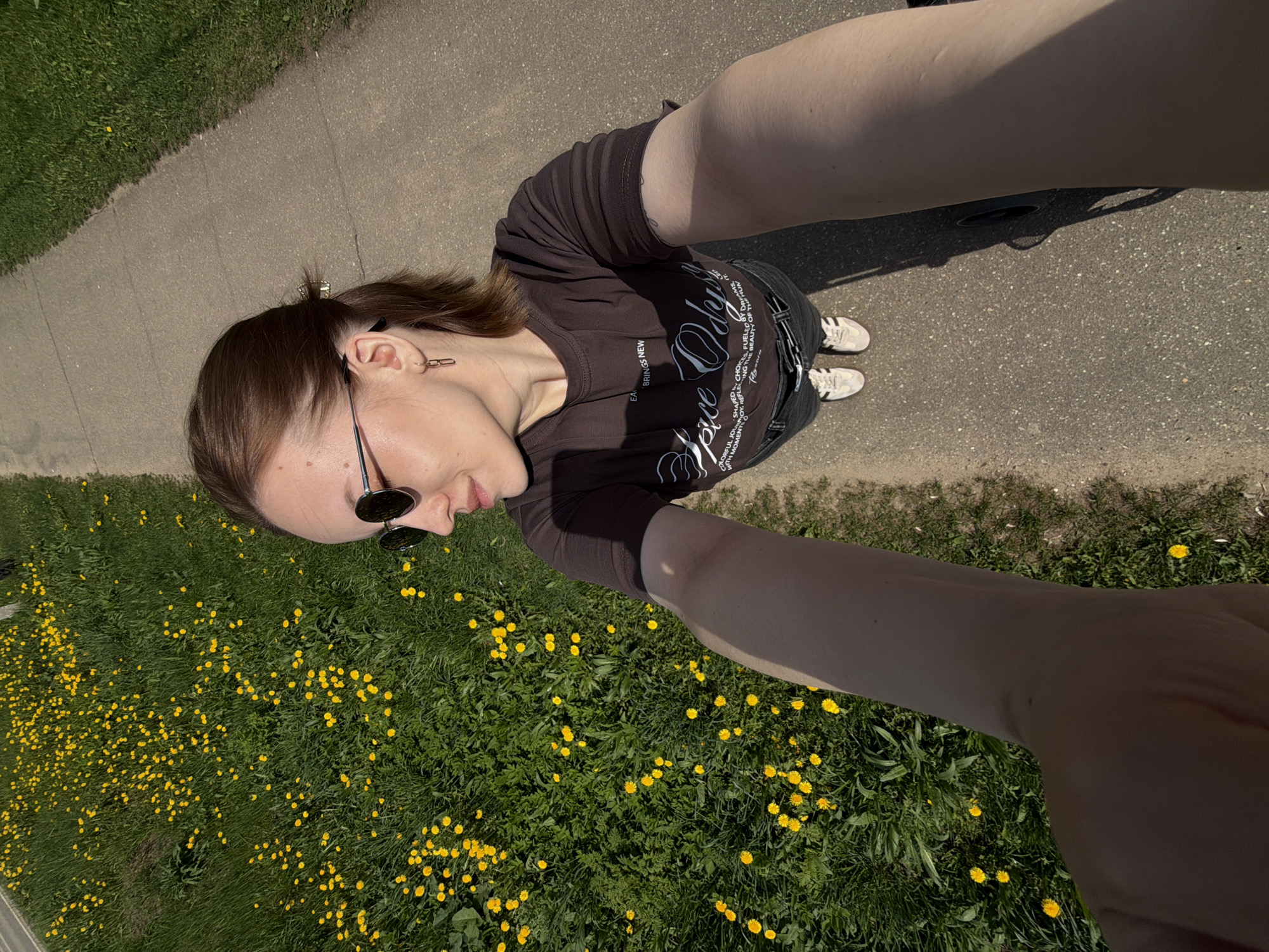 A young woman in a brown t-shirt and jeans stands on a paved path near a grassy field dotted with yellow dandelions. She is wearing sunglasses and earrings. Her clothing is slightly tight. She is taking a selfie from an overhead perspective. Her sneakers are white with black stripes. The location appears to be Reshetnikovo, Russia based on possible metadata (not directly visible) associated with the image.