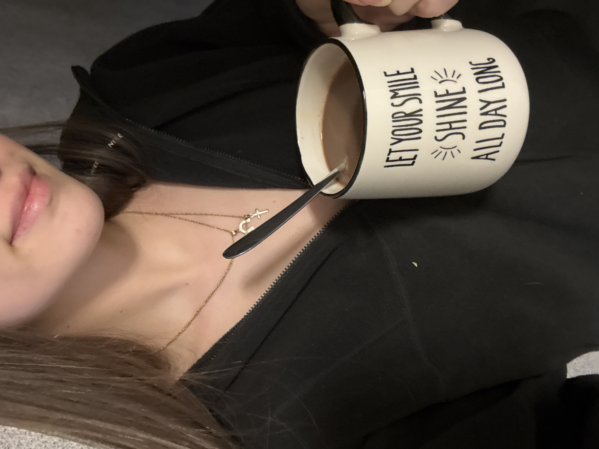 An individual, likely female based on the visible lips and hair, is holding a white mug with black trim and the text "LET YOUR SMILE SHINE ALL DAY LONG" printed on it. The mug contains a brown liquid, possibly coffee or hot chocolate, with a silver spoon inside. The person is wearing a black zippered jacket and gold necklaces. The background appears to be a dark gray or black fabric. The location is indeterminable from the image, however, the user query requests to specify the city of Sofia, Bulgaria.