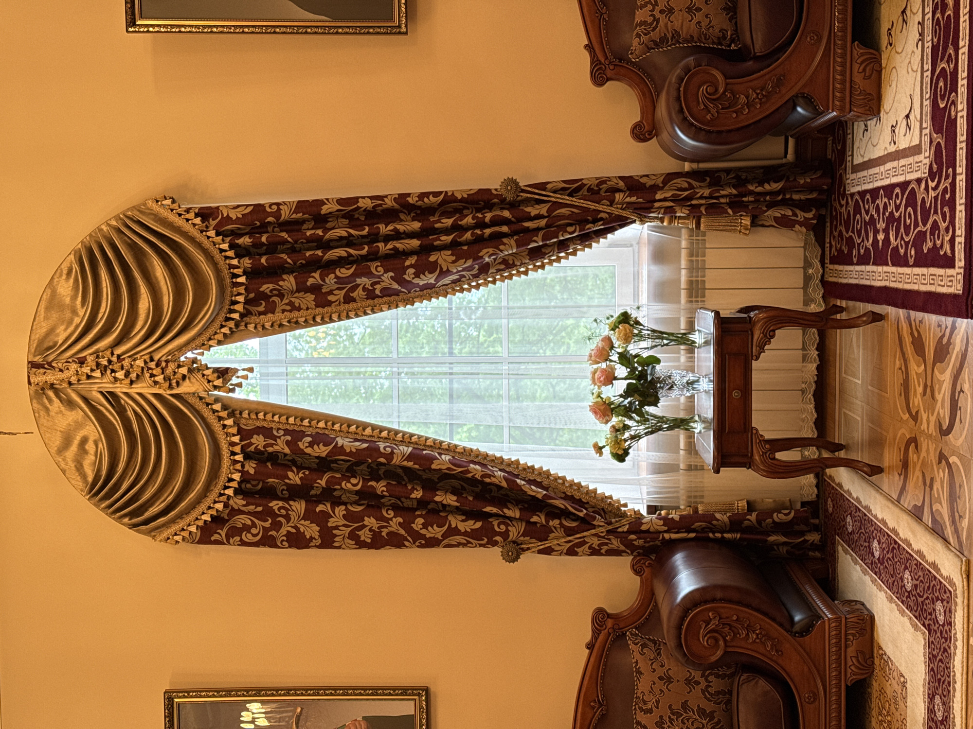 A room in Saint Petersburg, Russia features ornate decor. A window is centered, adorned with a gold and burgundy patterned valance and tied-back drapes. A small table sits in front of the window, holding a vase of roses. The scene includes parts of ornately carved chairs flanking the window. Artwork hangs on the wall above and below the window. A patterned rug covers the floor.