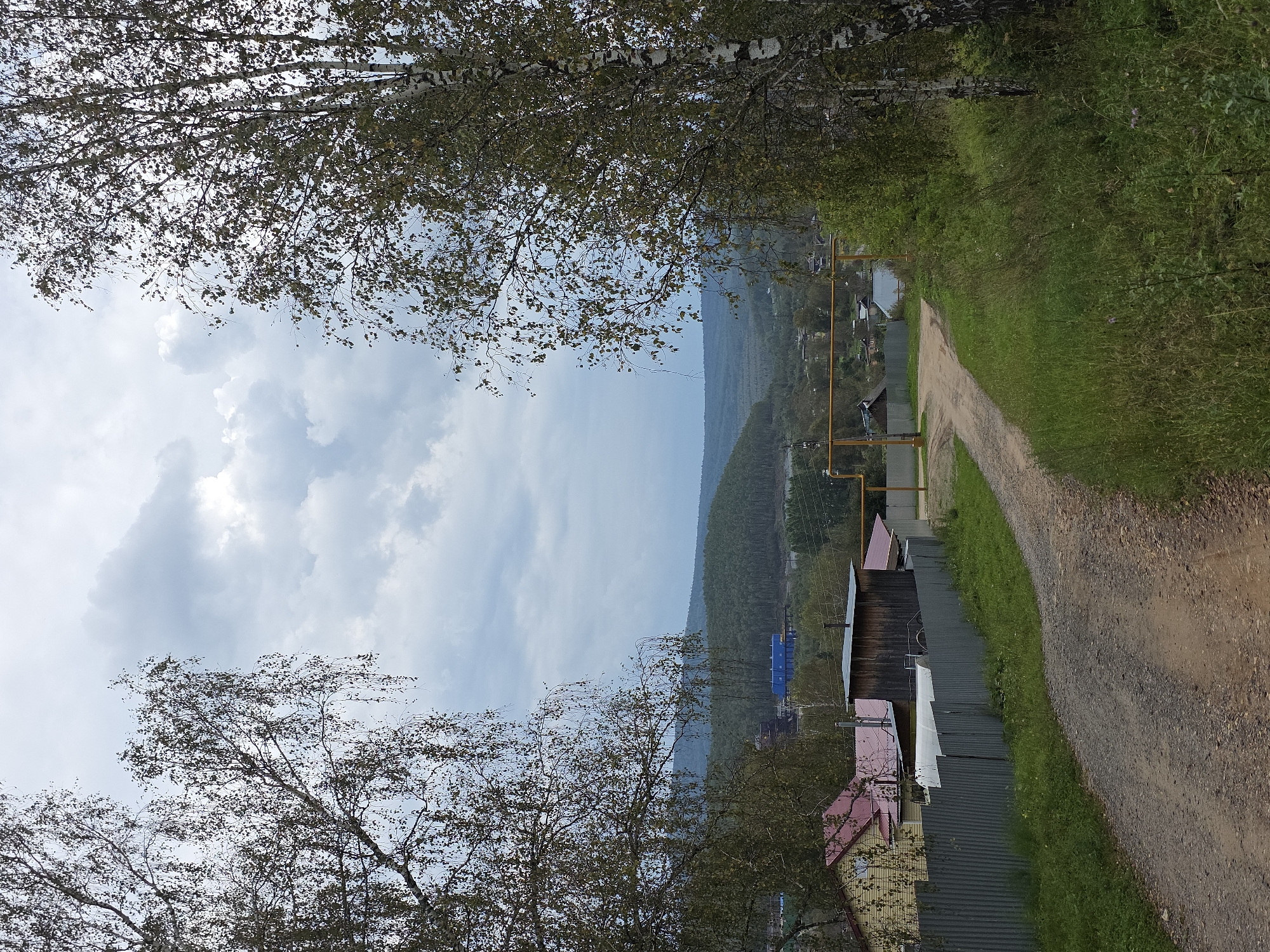 An overcast, late autumn day in Magnitka, Russia, is depicted in this image. The foreground is dominated by a gravel road leading into a village. To the left of the road, a grey corrugated metal fence encloses a property with a yellow house with a red roof and a dark wooden structure with a green roof. A large white cylindrical tank is visible behind the fence. Tall birch trees frame the scene, with the one on the right being particularly prominent. In the middle ground, a yellow gas pipe runs horizontally, supported by poles. Further into the village, more houses with red roofs are scattered among trees. Beyond the village, a dense forest covers rolling hills, and in the far distance, industrial buildings with blue roofs are visible against a backdrop of more forested hills under a cloudy sky. The overall impression is one of a rural, industrial fringe in a temperate climate.