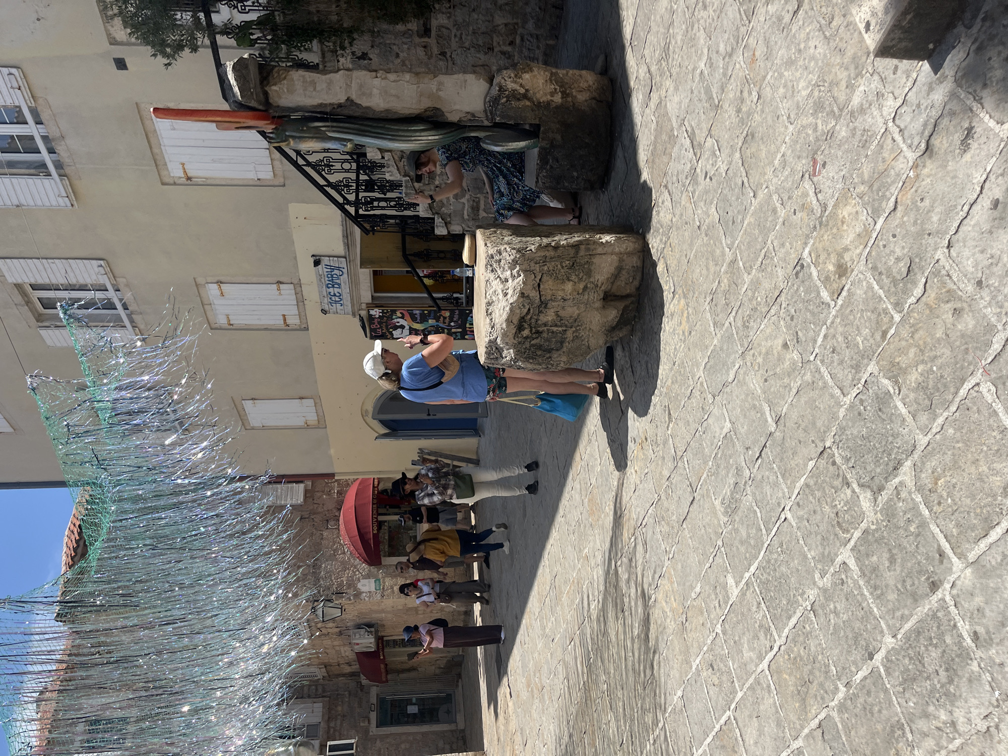 The image captures a bustling street scene in Budva, Montenegro. The setting features traditional stone buildings with closed white-shuttered windows. A decorative net-like installation hangs overhead. The street is paved with cobblestones. A group of pedestrians are present, including a woman in a blue shirt and a white cap standing near a shop with a sign reading "Ice Baby". Another woman is interacting with what appears to be a decorative sculpture attached to the wall. Shops with red awnings are visible.